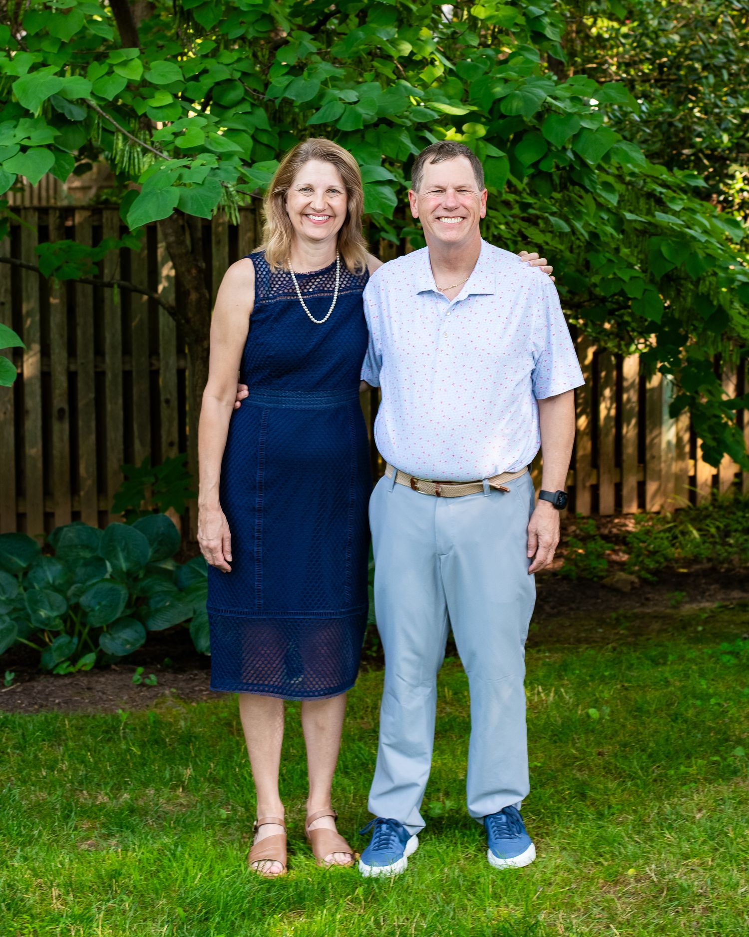 A smiling couple poses together on a green lawn with lush garden foliage and wooden fence backdrop.