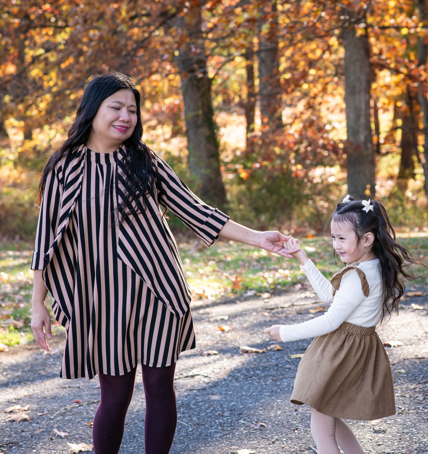A mother and daughter hold hands and dance together outdoors among colorful autumn trees.