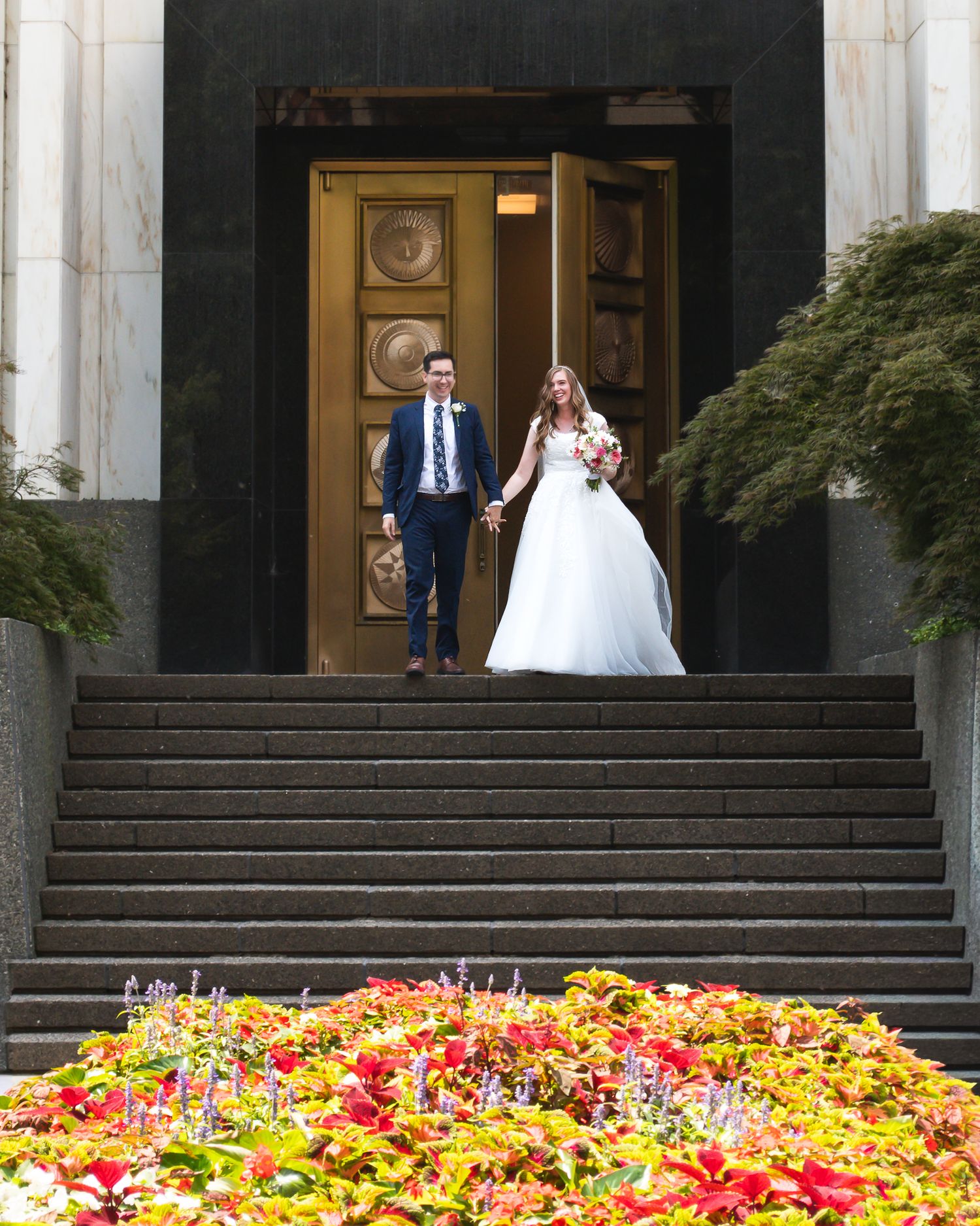 Bride and groom holding hands on grand stone steps with vibrant red and yellow flowers in foreground.