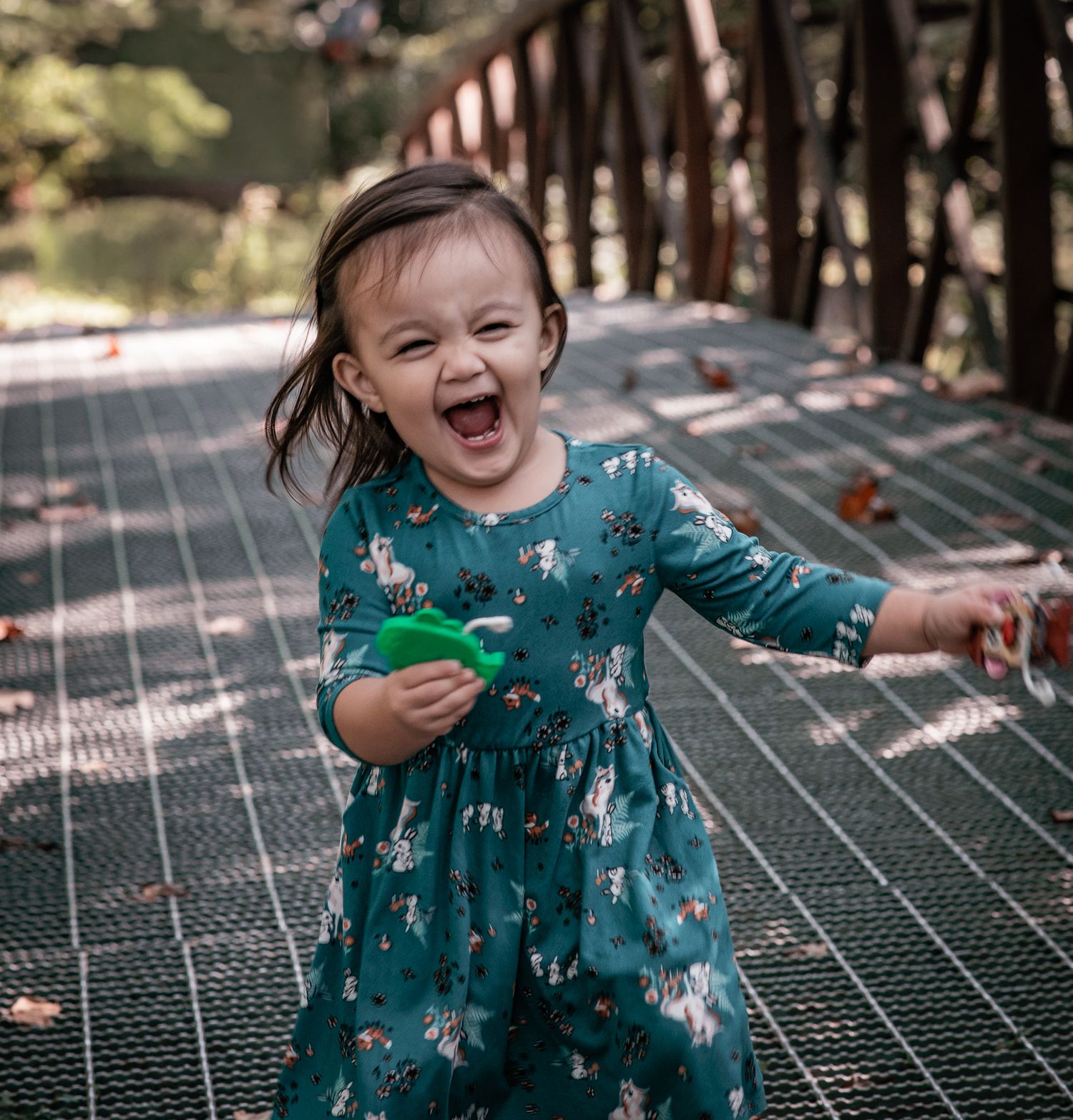 A joyful toddler girl in a teal floral dress laughs while playing on a metal bridge outdoors.