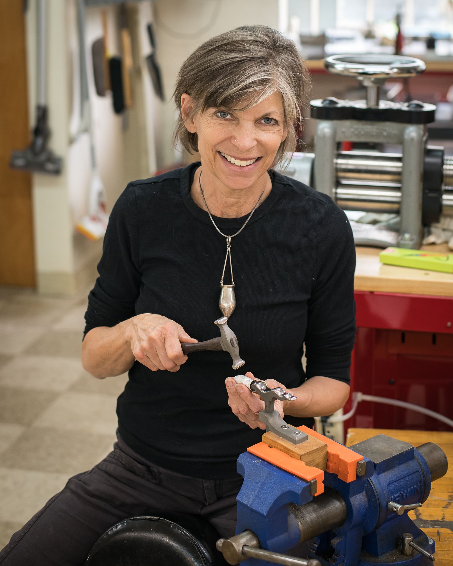 Smiling woman with gray hair uses a hammer at a workbench vise in a jewelry or metalworking studio.