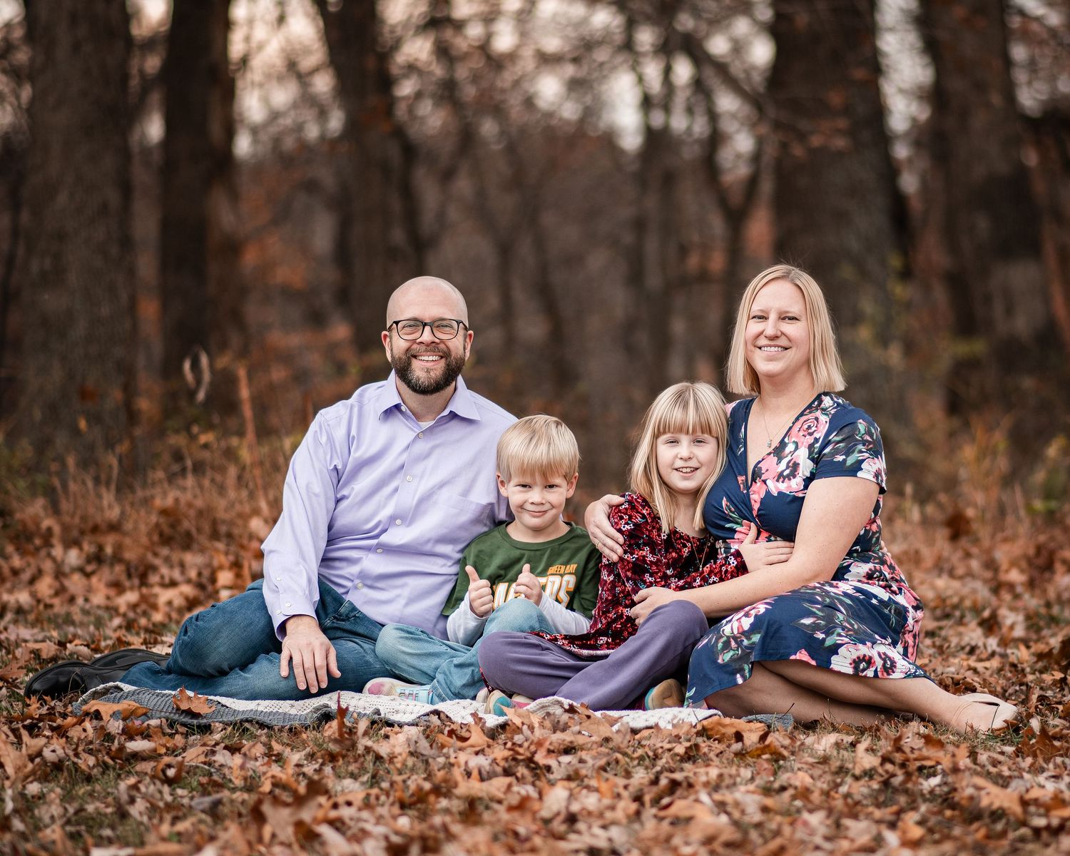 A family lounges together on autumn leaves in a wooded setting during an outdoor photo session.