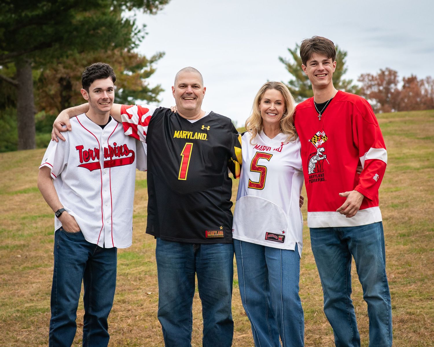 Family of four posing outdoors in Maryland Terrapins sports jerseys, smiling together on a grassy field.