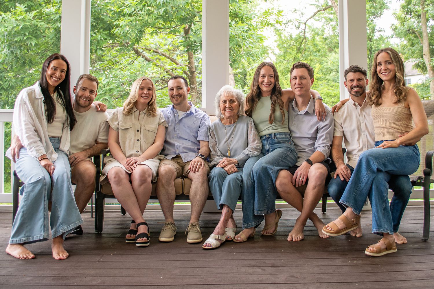 Multi-generational family smiling together on a covered porch surrounded by lush green trees.
