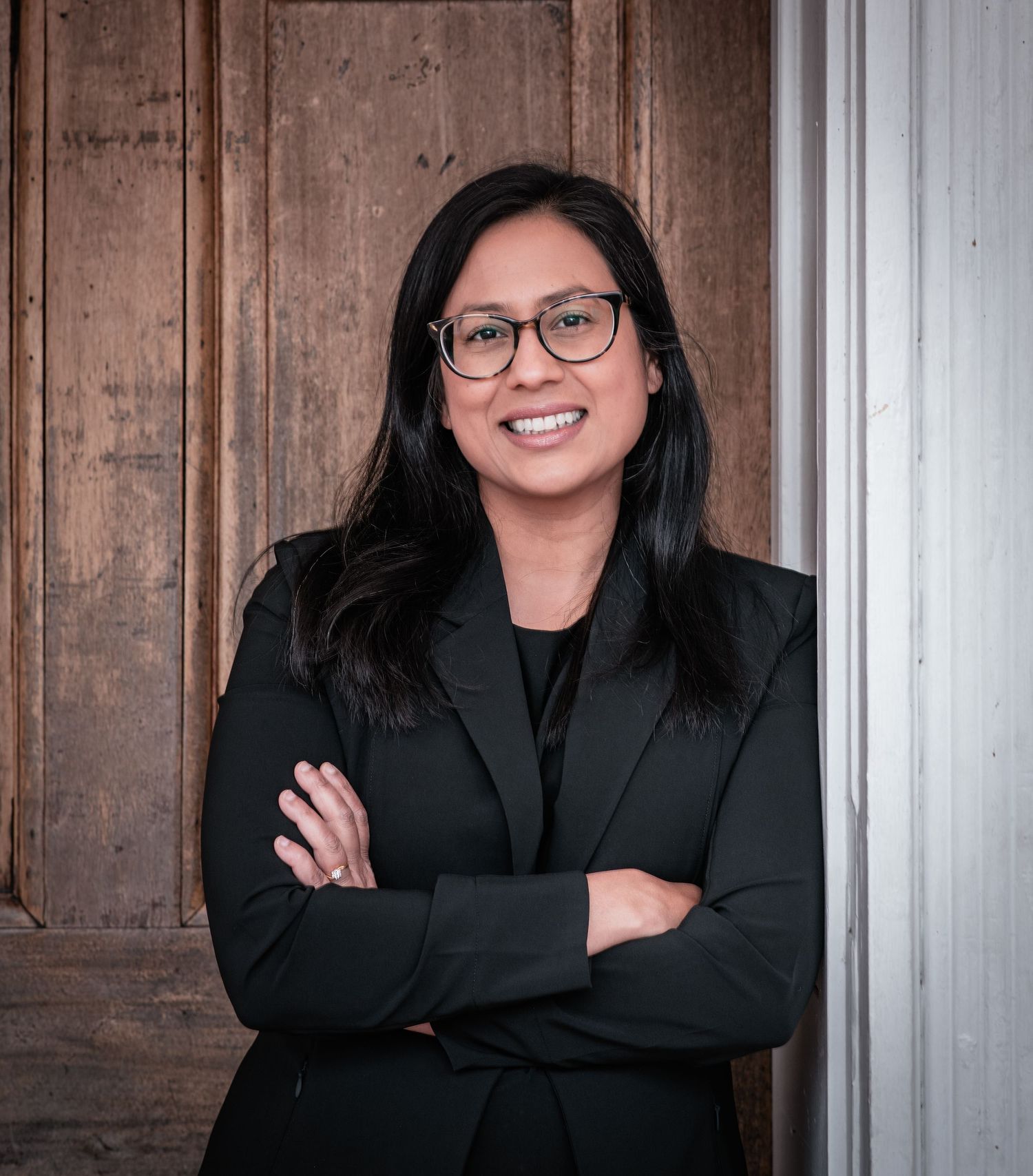 Professional headshot of a person in a black blazer smiling warmly against a wooden paneled wall.