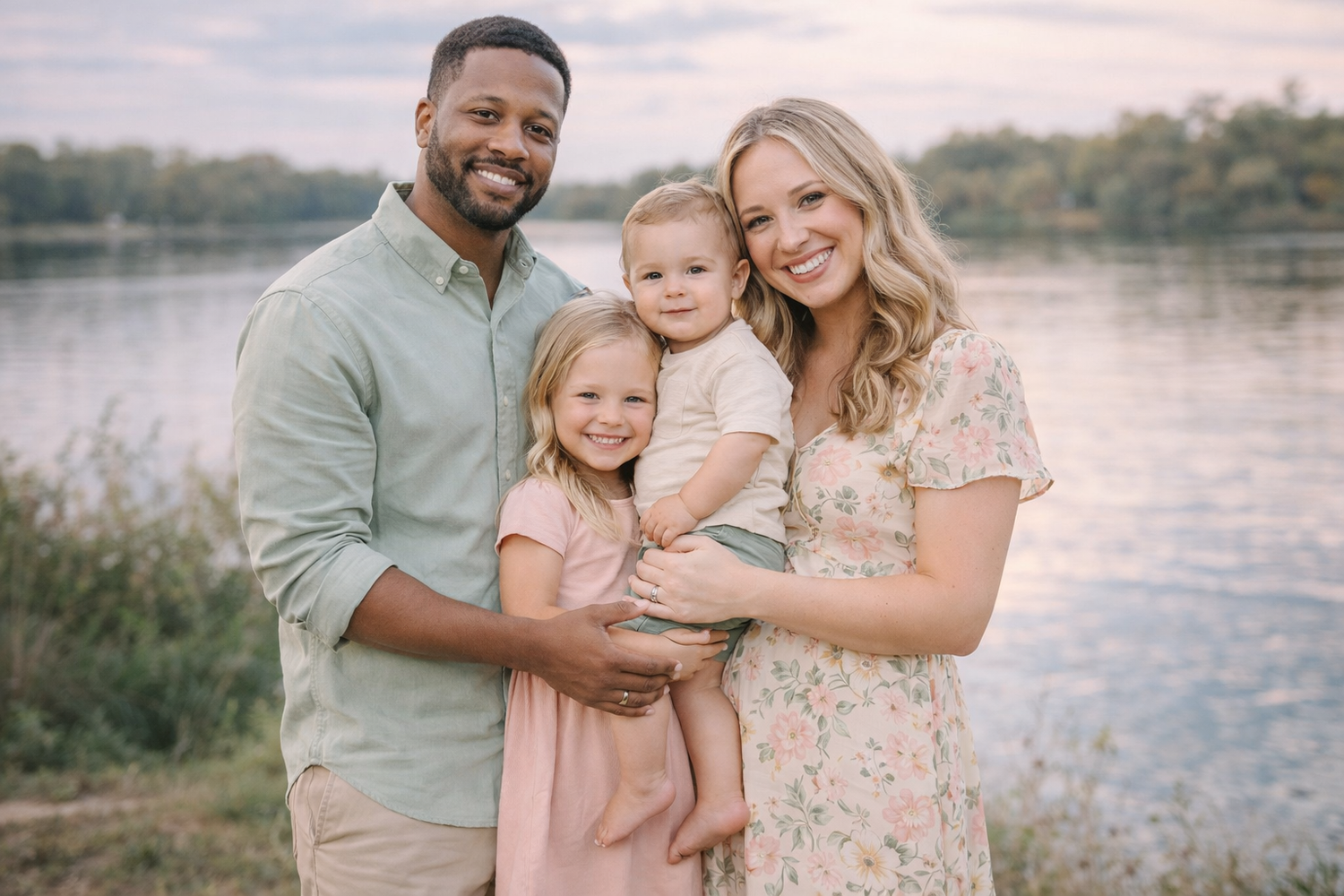 A family of four stands together smiling by a peaceful lake at sunset wearing coordinated pastel outfits.