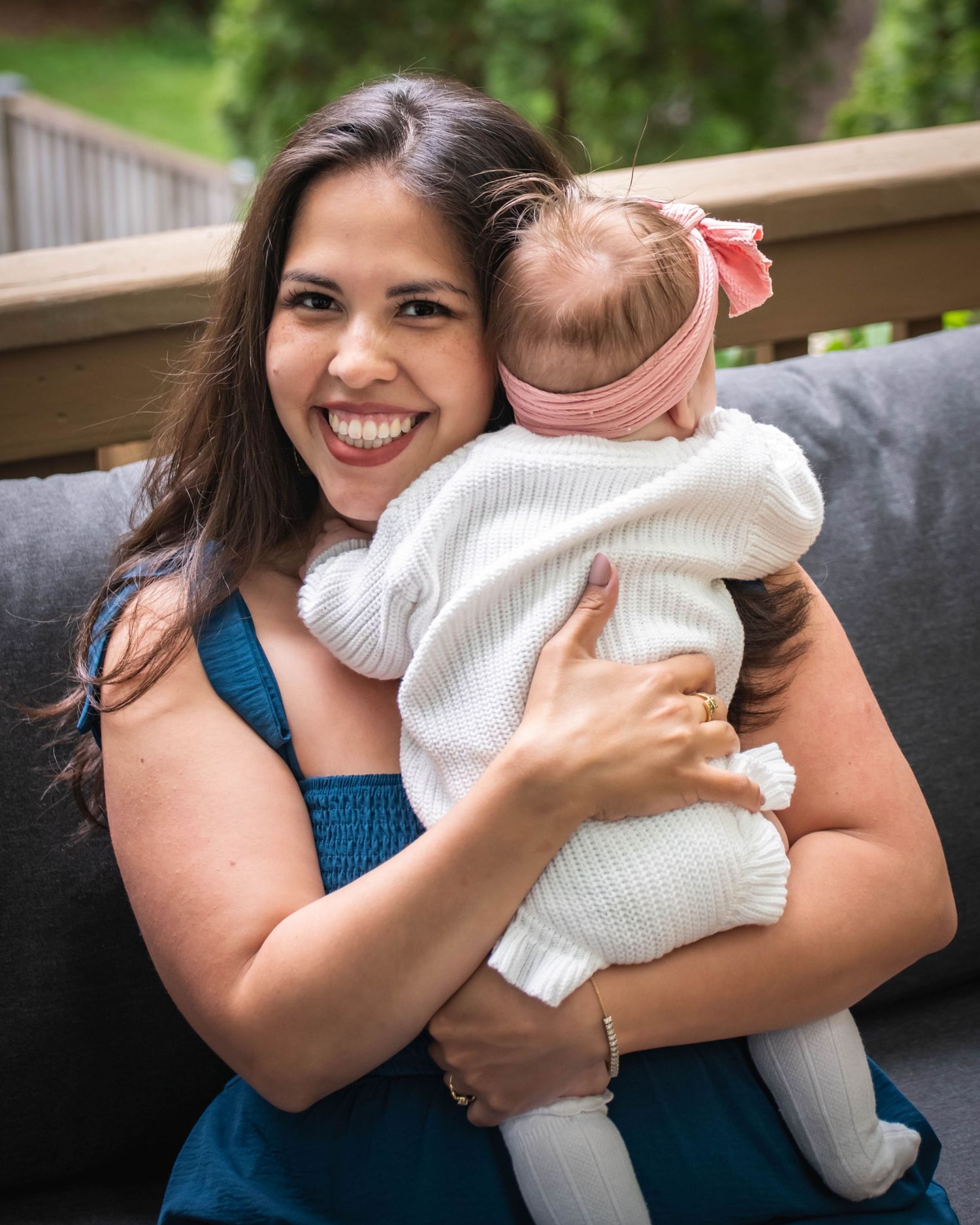 Smiling woman in teal dress hugging a baby girl wearing white knit outfit and pink bow headband outdoors.