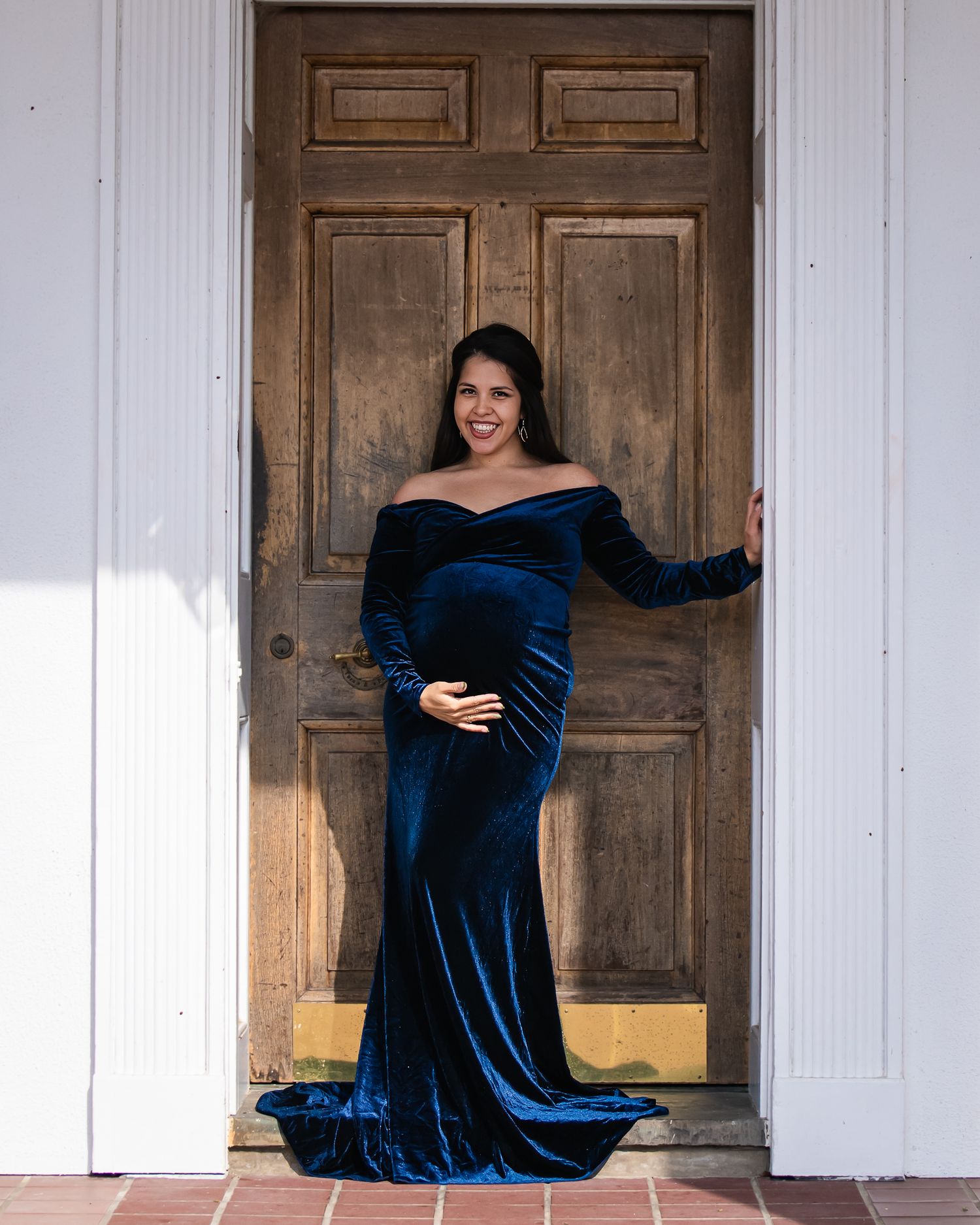 Pregnant woman in elegant navy blue velvet off-shoulder gown posing in front of rustic wooden door.