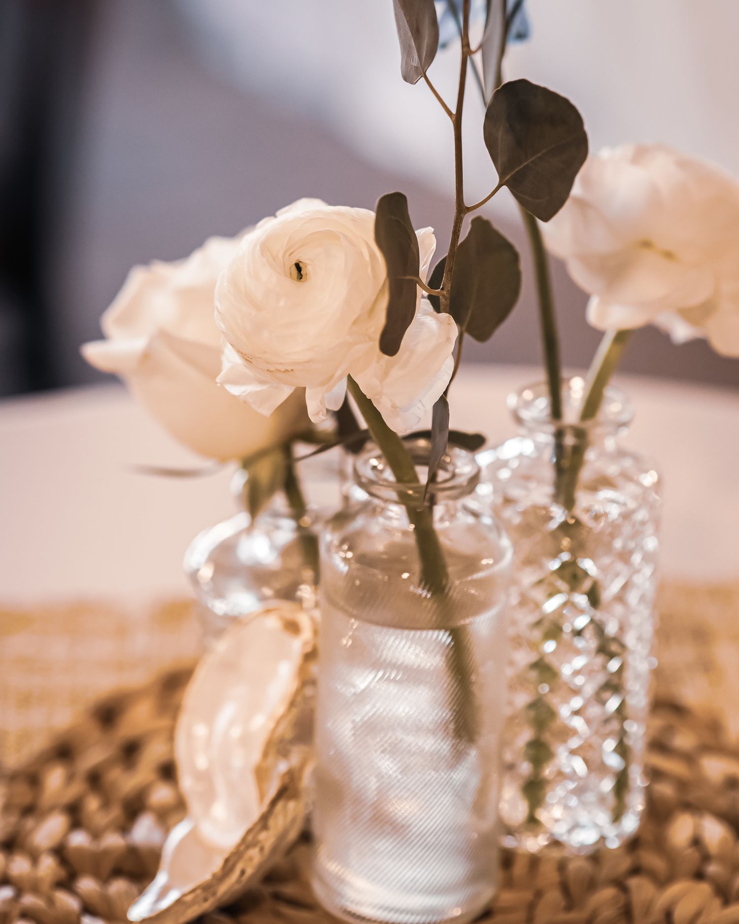 White ranunculus and roses in vintage glass bud vases on a woven mat with an oyster shell accent.