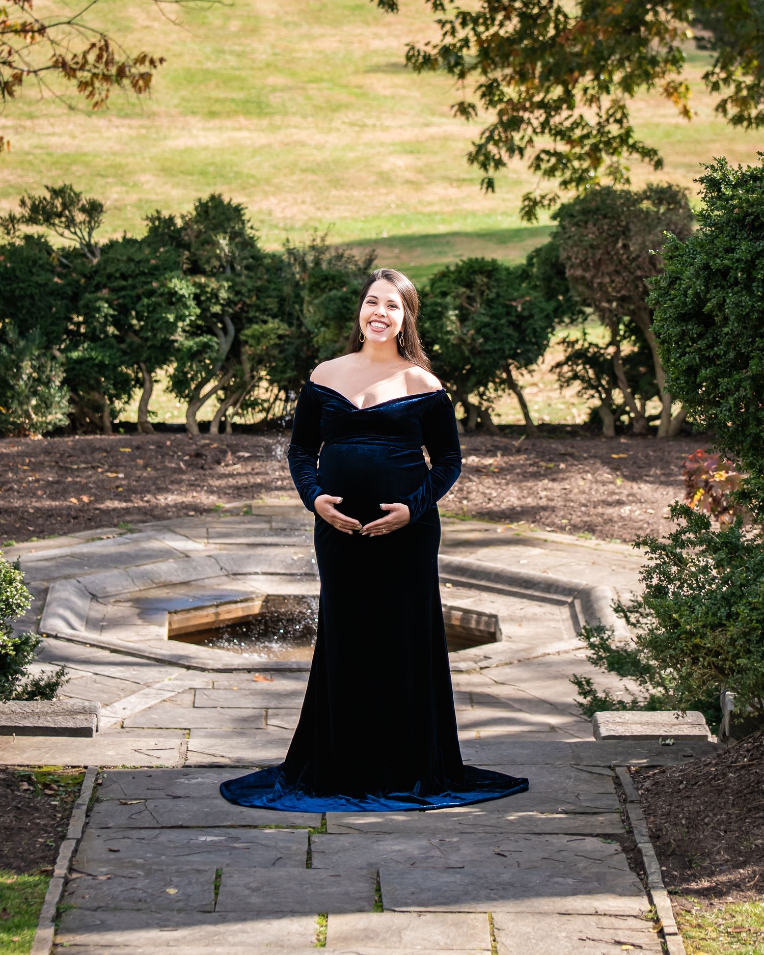 Pregnant woman in elegant navy blue velvet off-shoulder gown posing in a garden for a maternity photo shoot.