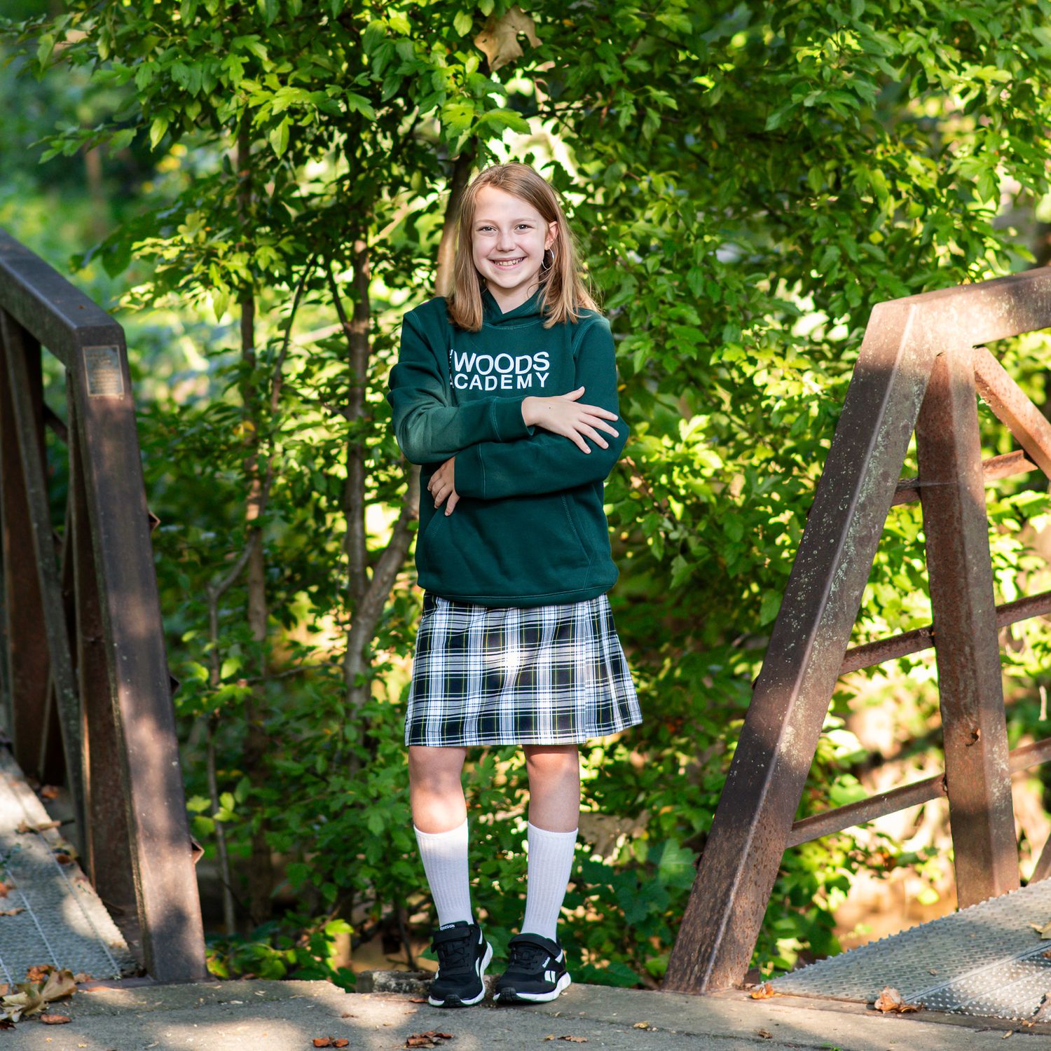 Girl in Woods Academy green sweatshirt and plaid skirt stands smiling on a wooden bridge outdoors.