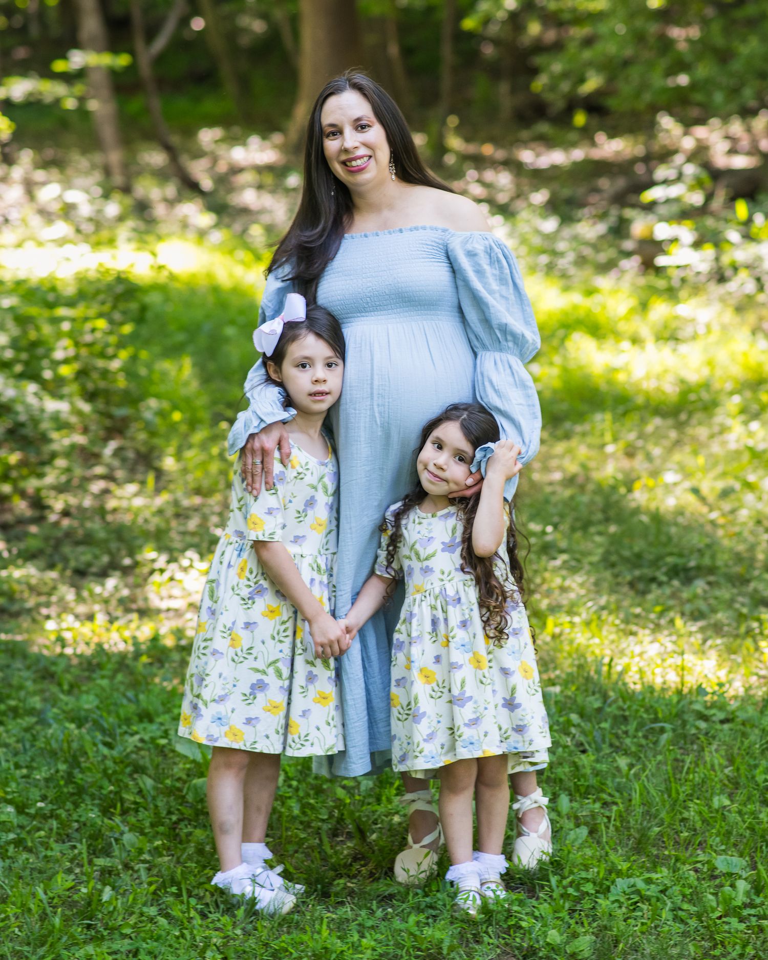 Pregnant woman in blue dress poses outdoors with two young girls in matching floral dresses in a lush green setting.