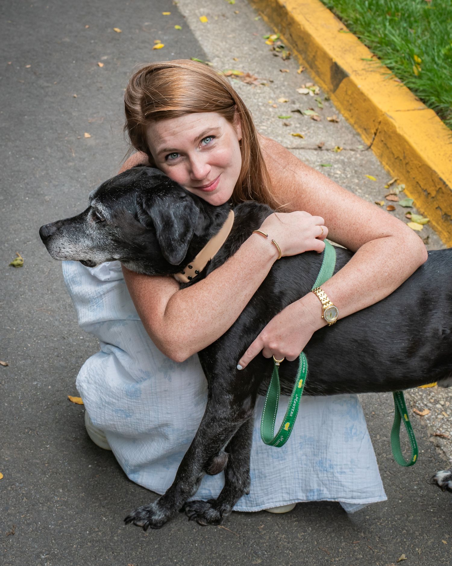 Woman with brown hair hugging a black dog on a leash outdoors on a paved path.