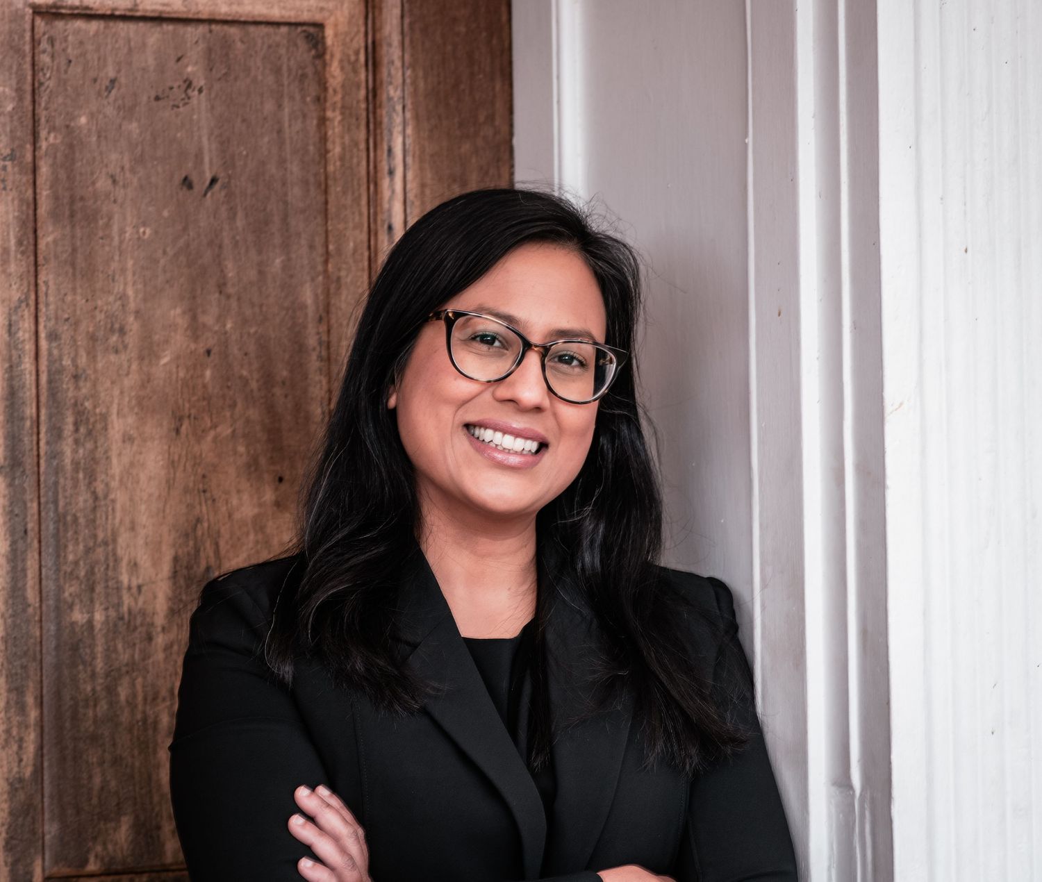 A person with long dark hair and glasses smiles while wearing a black top against a wooden wall background.