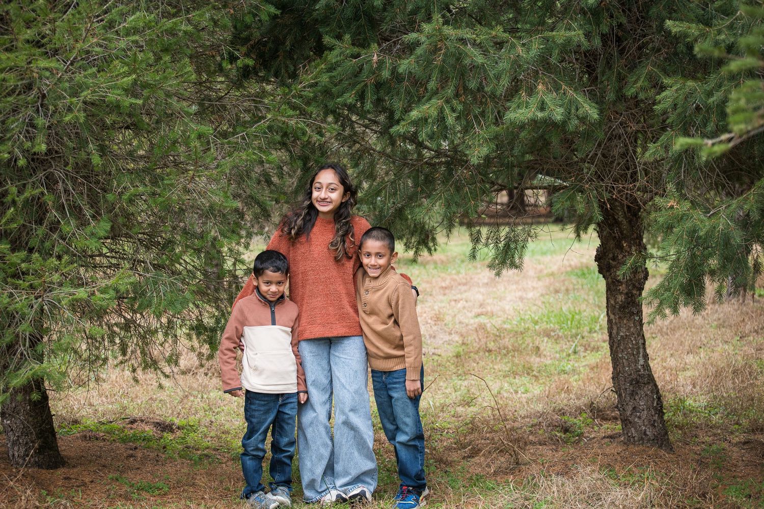 A family poses together in casual attire among evergreen trees in a natural outdoor setting.