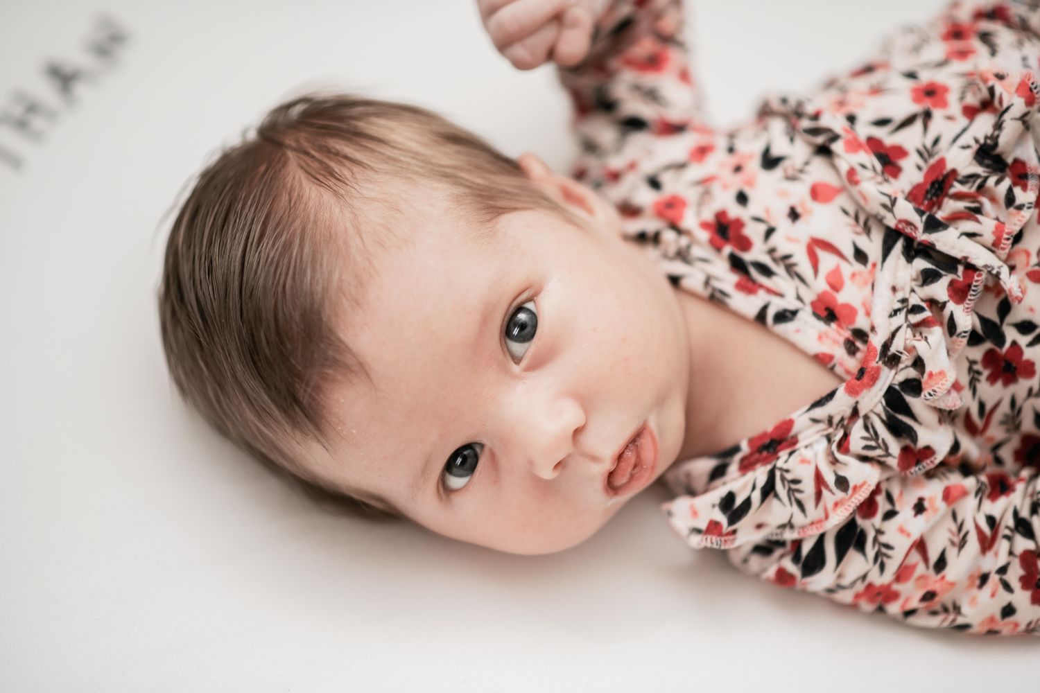 A baby lies on white bedding while wrapped in a floral swaddle blanket with red and black pattern.