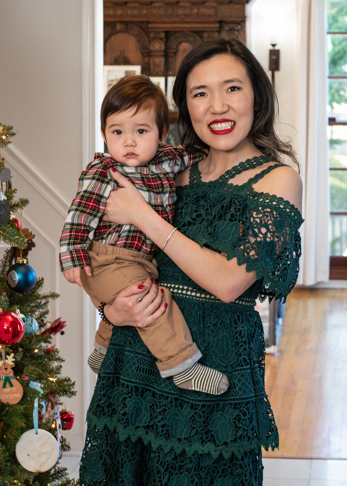 Mother and child in matching holiday outfits pose together in front of decorated Christmas tree in bright home interior.