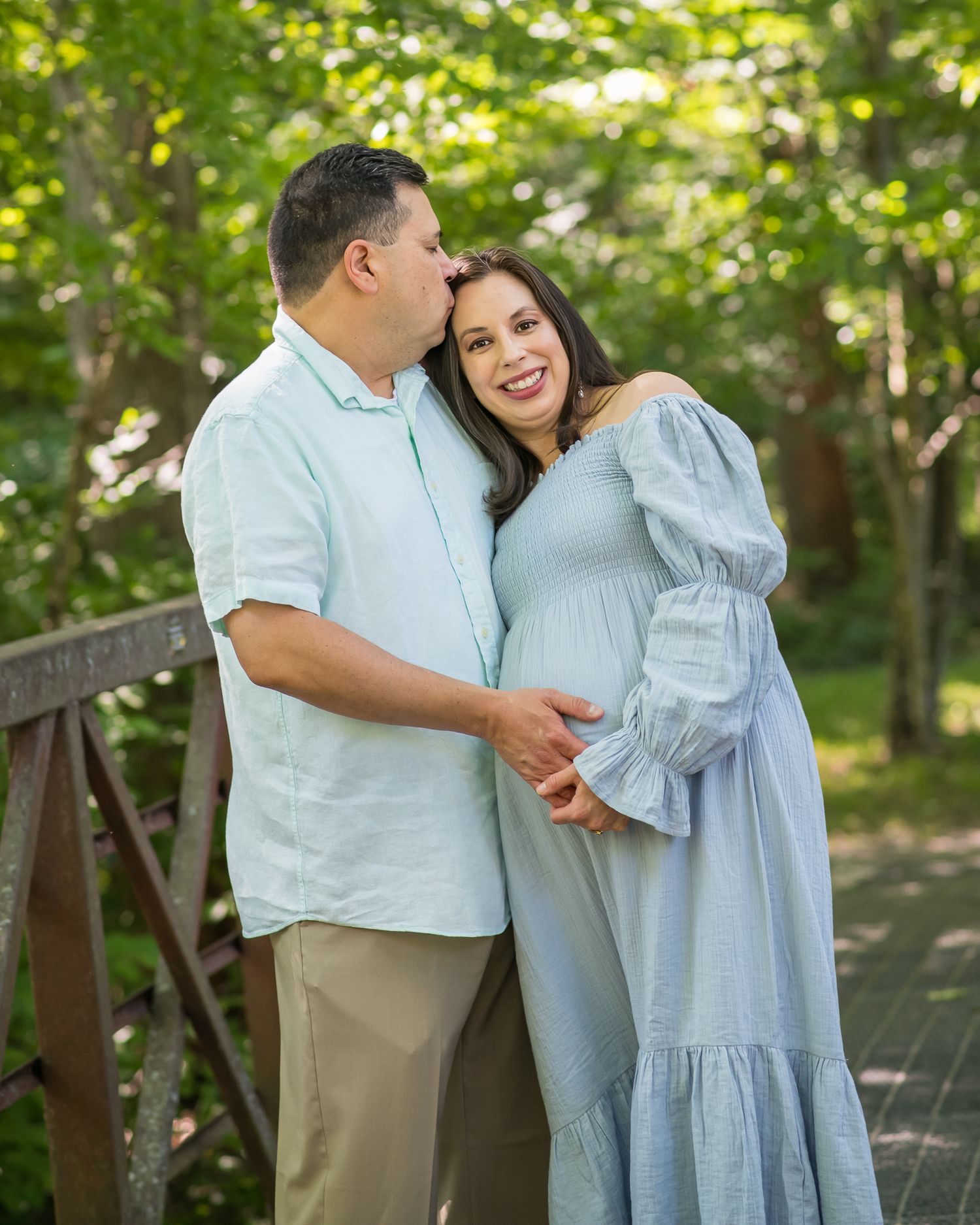 Couple poses on wooden bridge, man kisses pregnant woman in blue dress, surrounded by lush green trees.