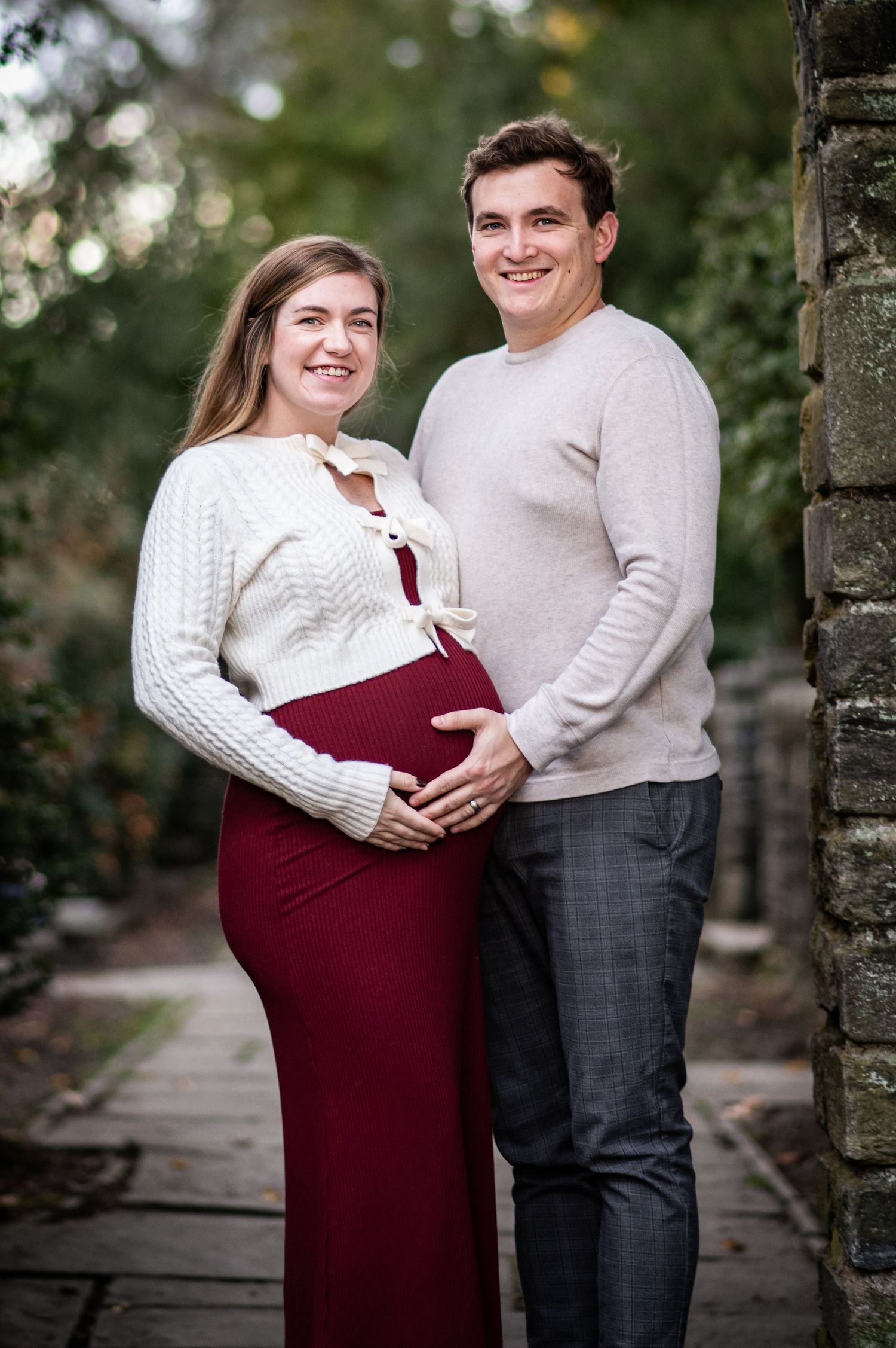 A couple in matching white sweaters embrace outdoors while showing off a burgundy maternity dress.