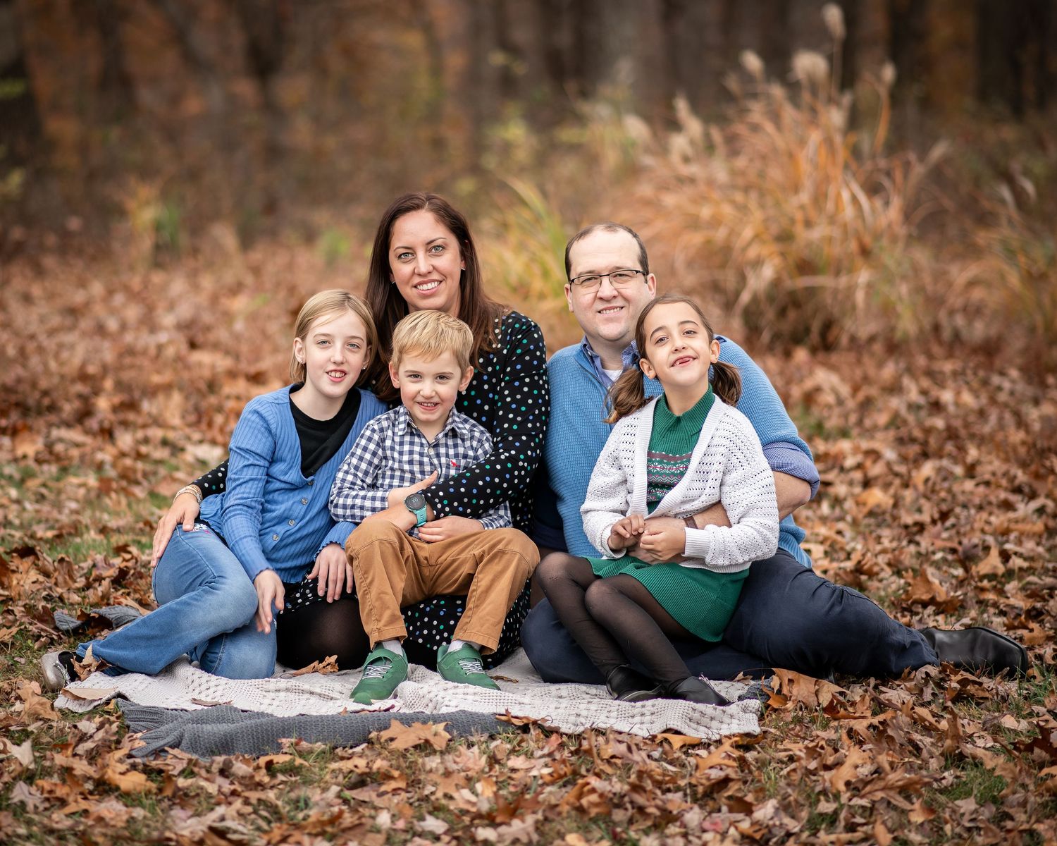 Family of five smiling together outdoors in autumn, sitting on a blanket surrounded by fallen leaves.