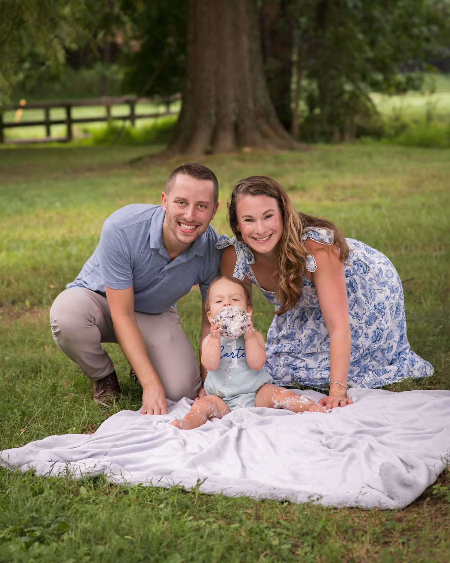 A smiling family poses on a white blanket outdoors, with parents flanking a baby eating cake near a large tree.