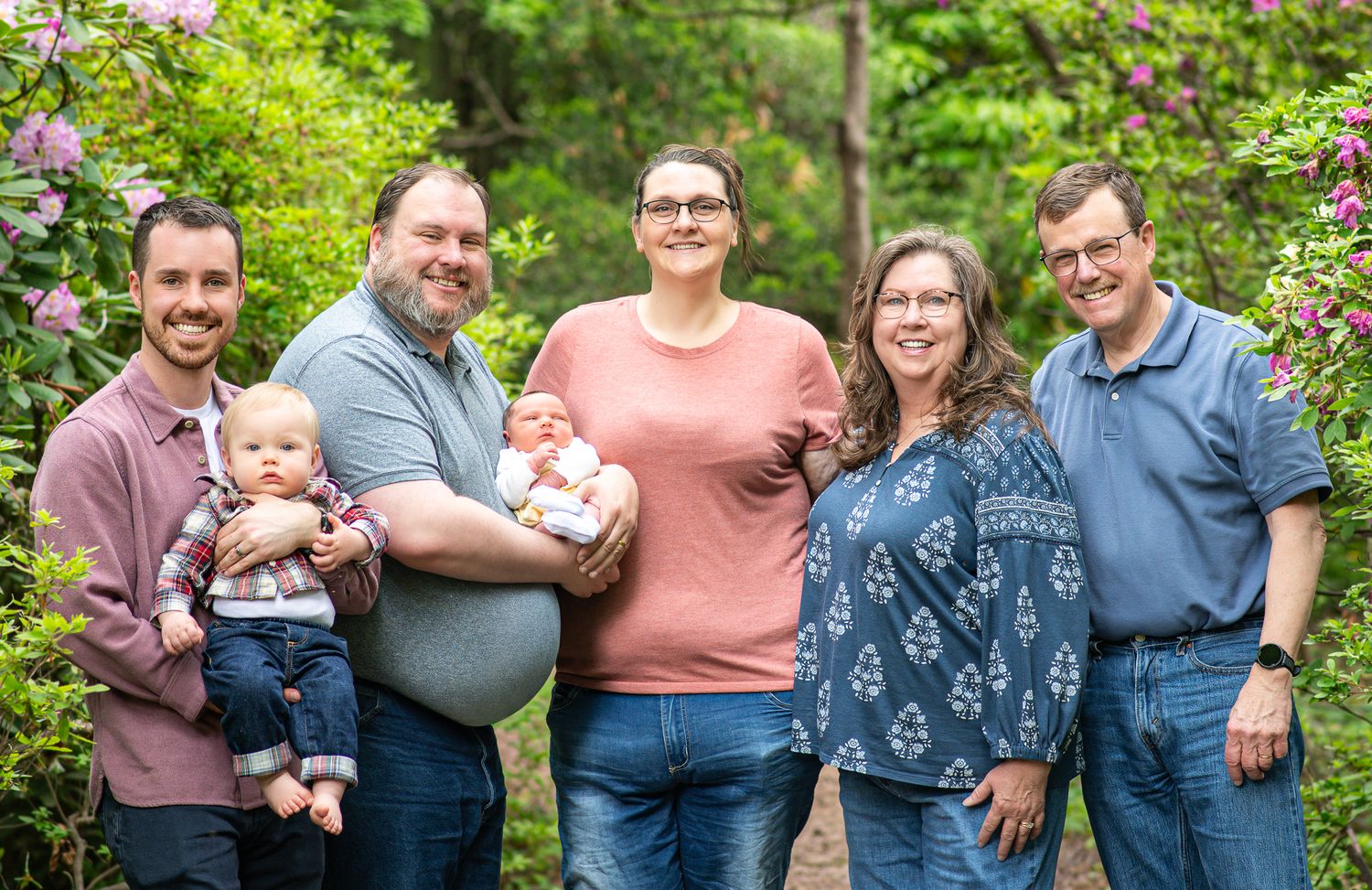 Family portrait of six adults with two babies outdoors among blooming pink flowers and lush greenery.