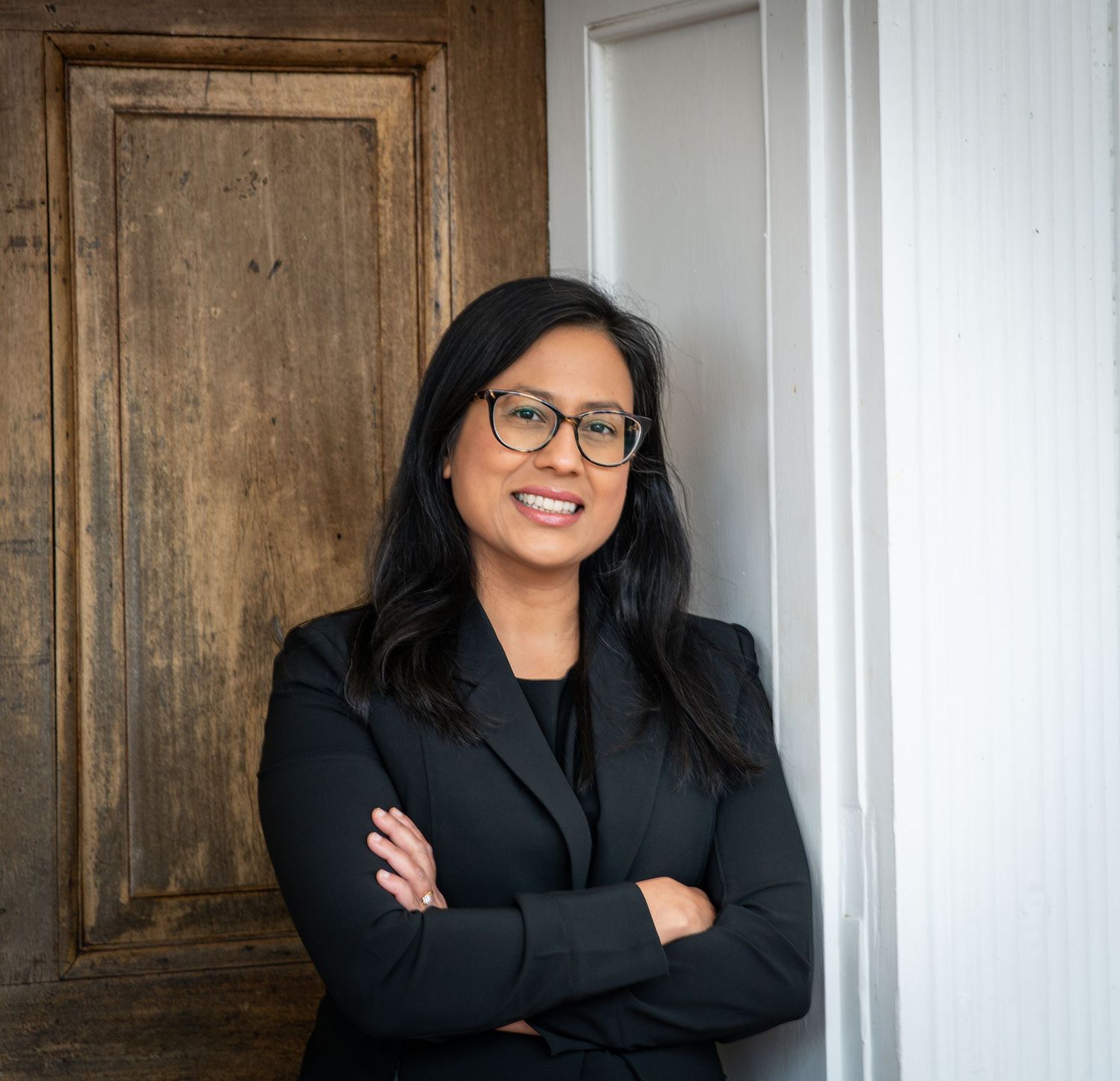 Professional wearing a black blazer and glasses stands confidently against a wooden door and white wall background.