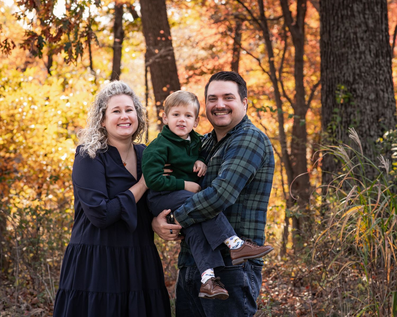 Family of three posing outdoors in autumn forest, parents holding toddler in green outfit among fall foliage.