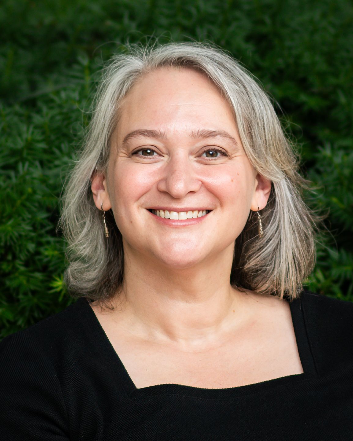 Smiling woman with gray hair wearing black top, posing outdoors with green foliage background.