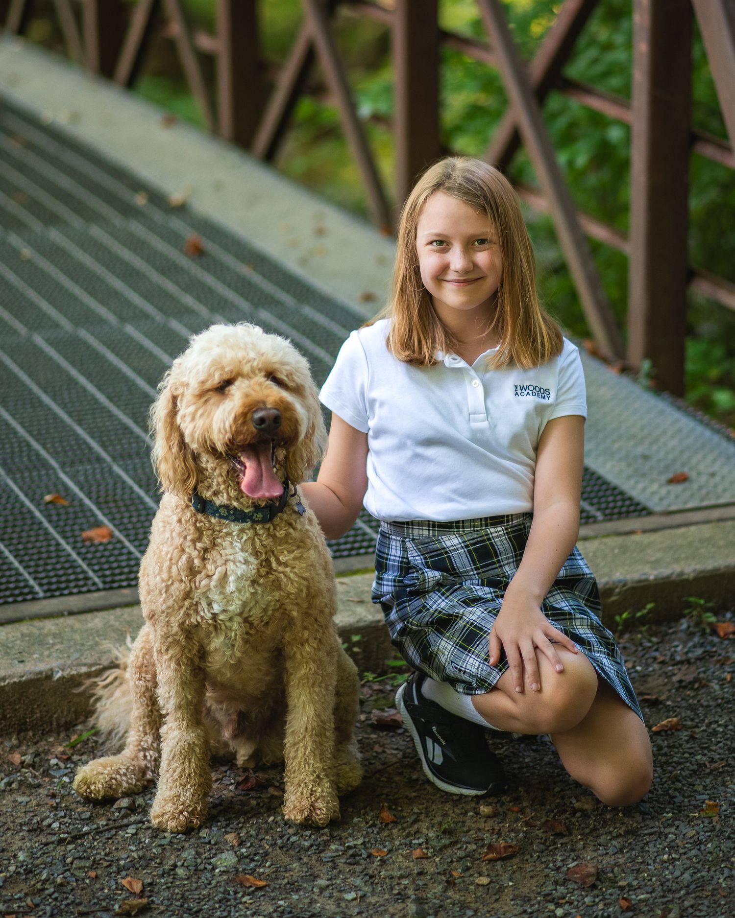 A smiling girl in school uniform kneels beside a fluffy golden doodle dog on a bridge path surrounded by greenery.