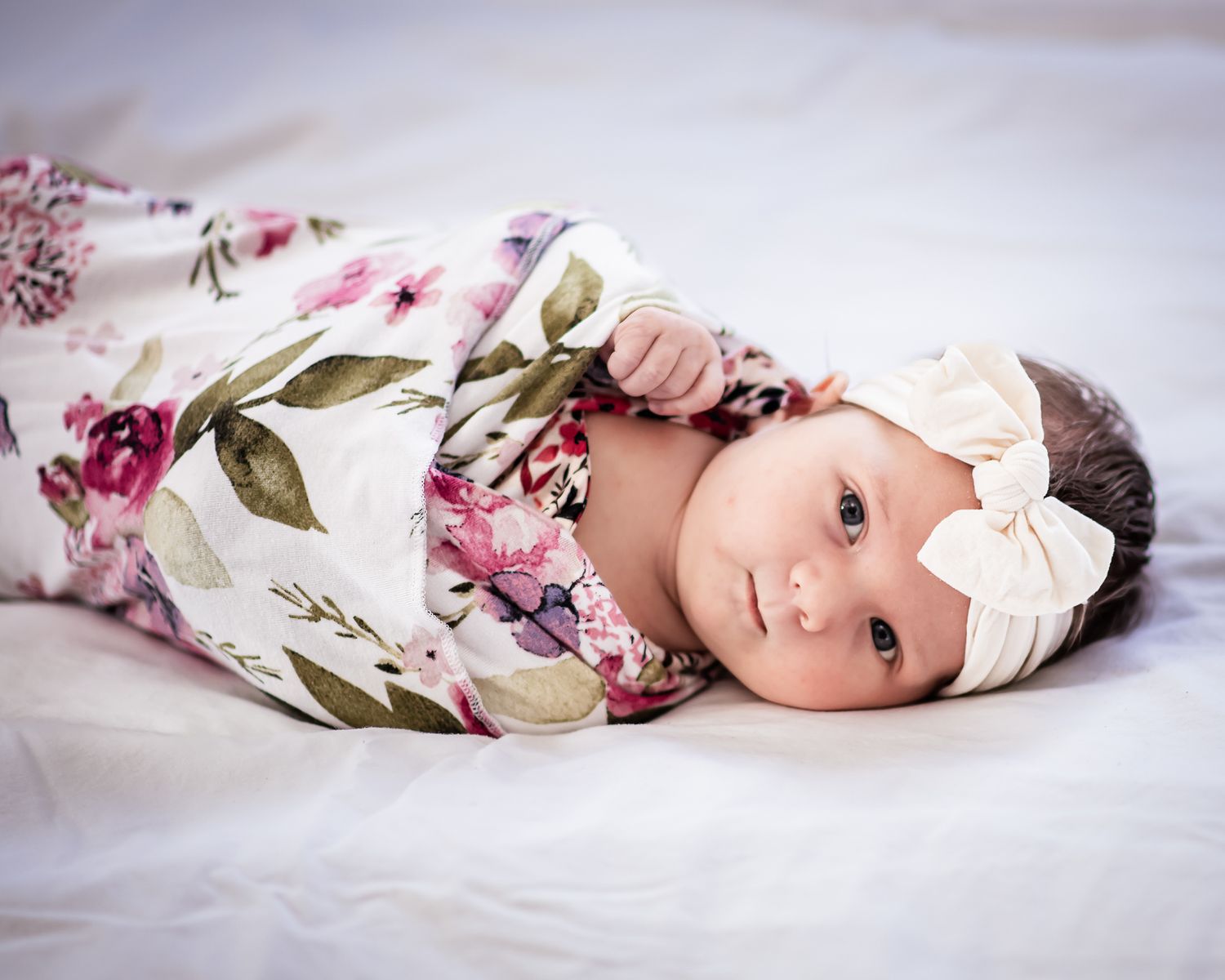 A baby wrapped in floral swaddle blanket lays on a soft white surface while looking at the camera.