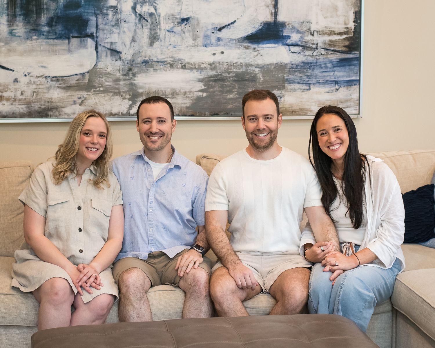 Four smiling adults sitting together on a couch in front of a modern abstract painting in a bright living room.