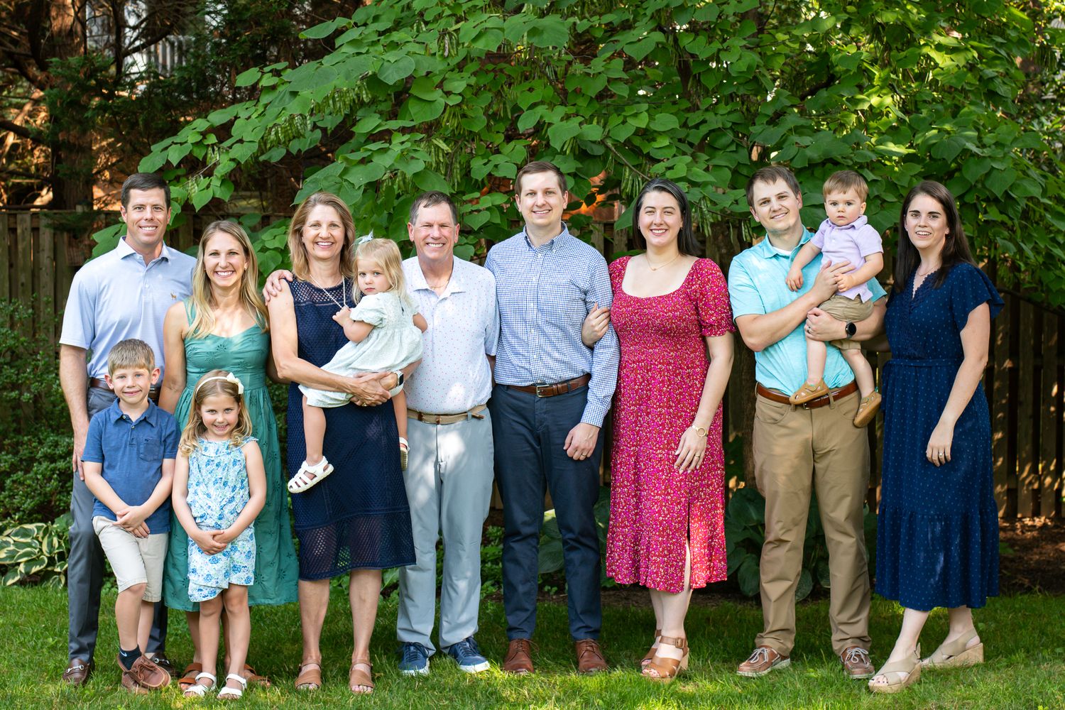 Large family group portrait smiling outdoors on green grass with lush garden background on a sunny day.