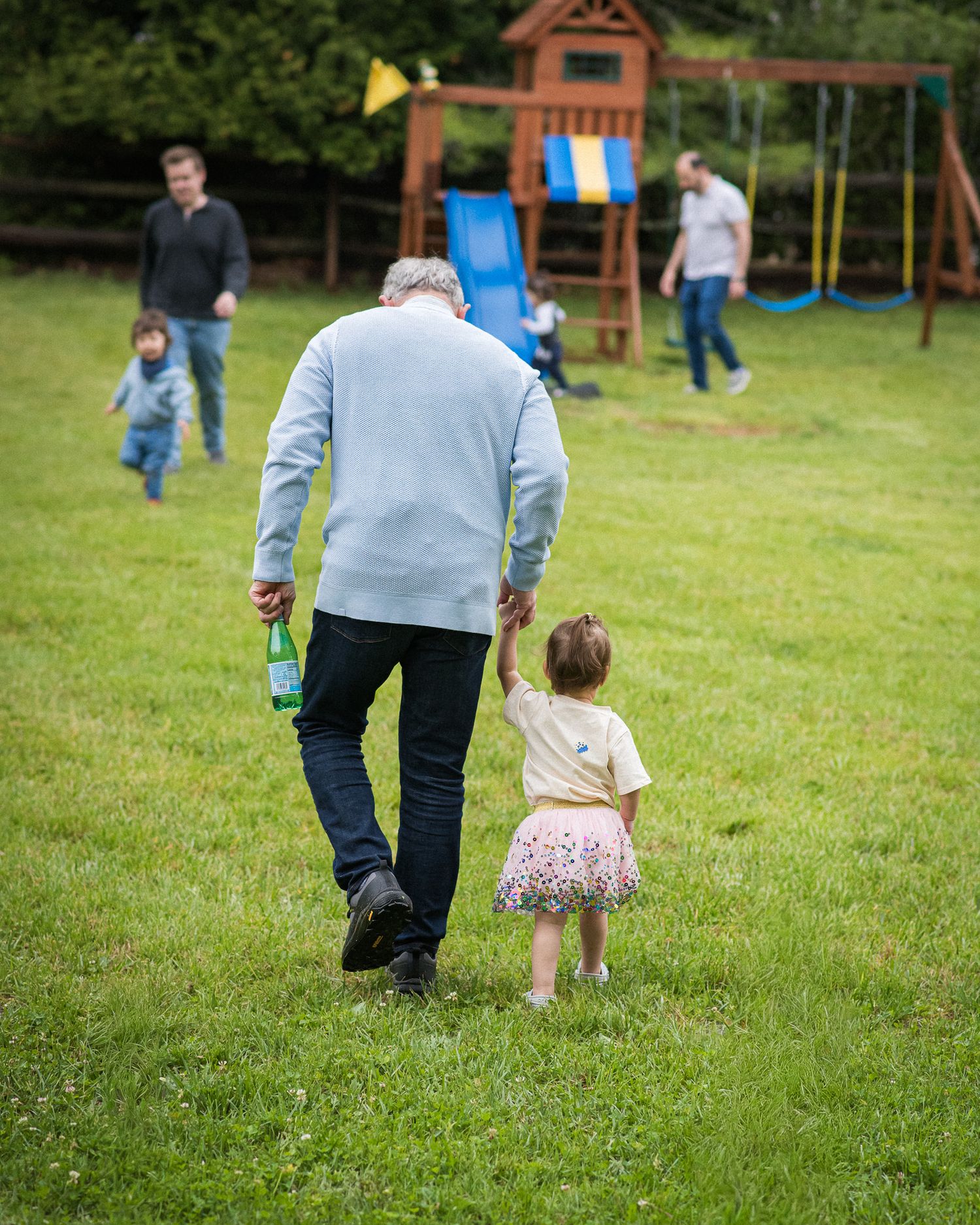 An elderly man holds a toddler's hand while walking across a green lawn toward a colorful playground.