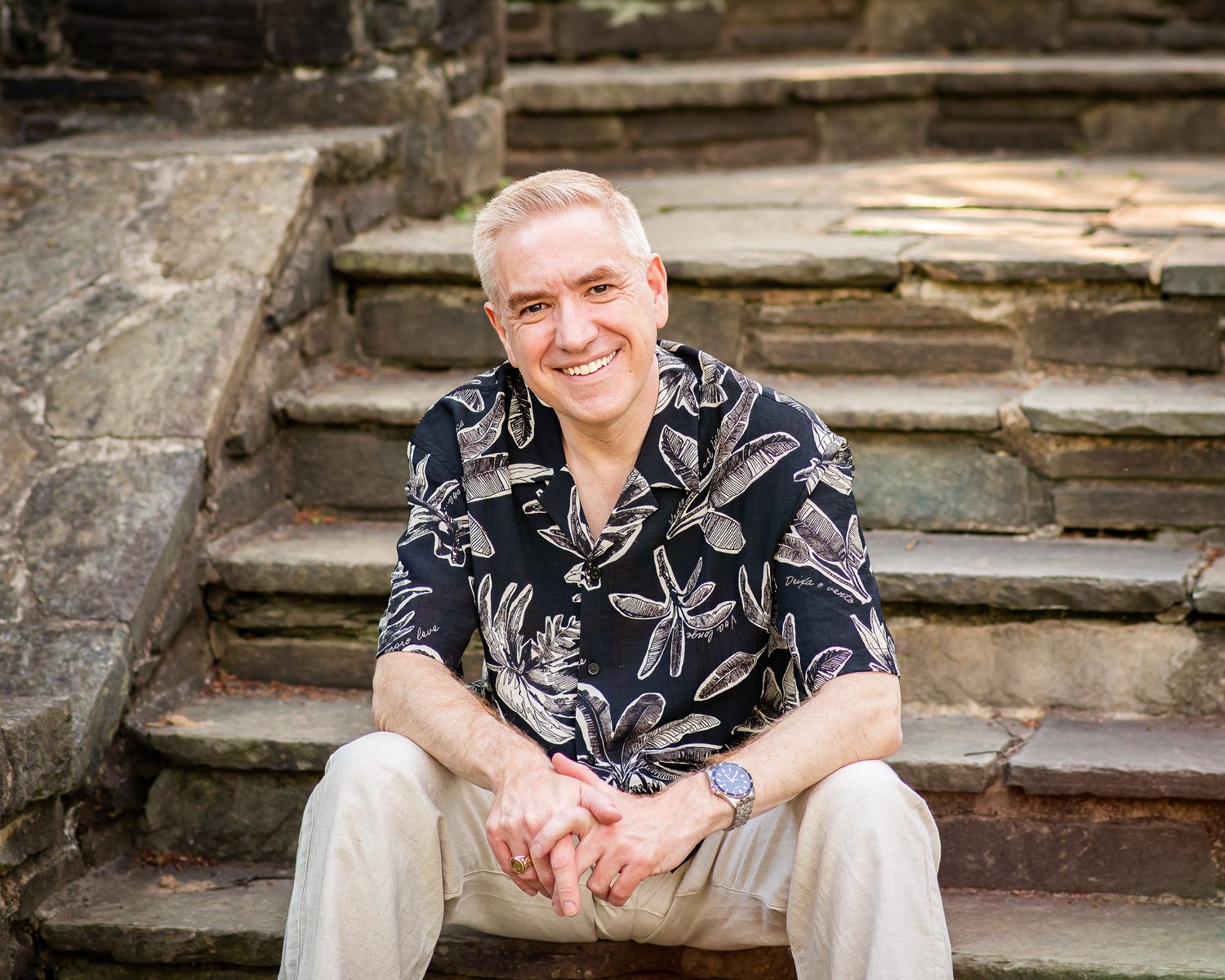 Smiling middle-aged man with gray hair sits on stone steps wearing a floral shirt and khaki pants outdoors.