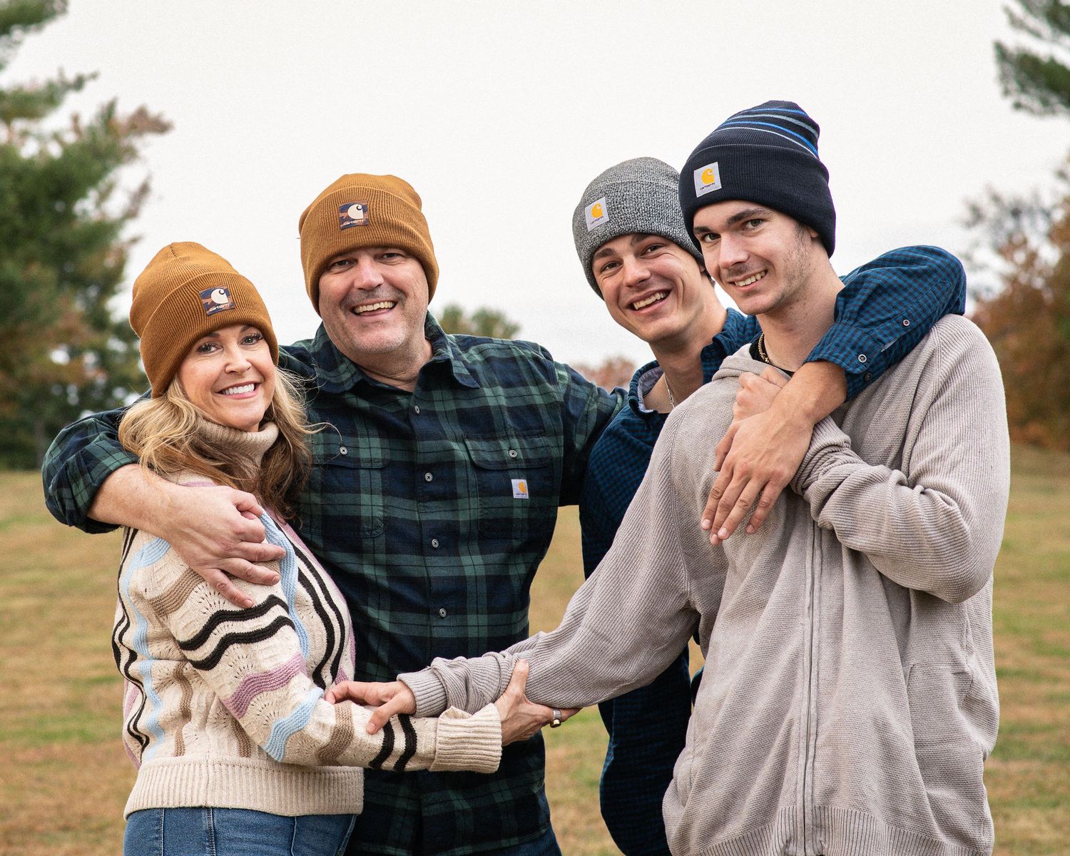 Four people wearing Carhartt beanies smile and embrace outdoors in a fall setting with trees in the background.