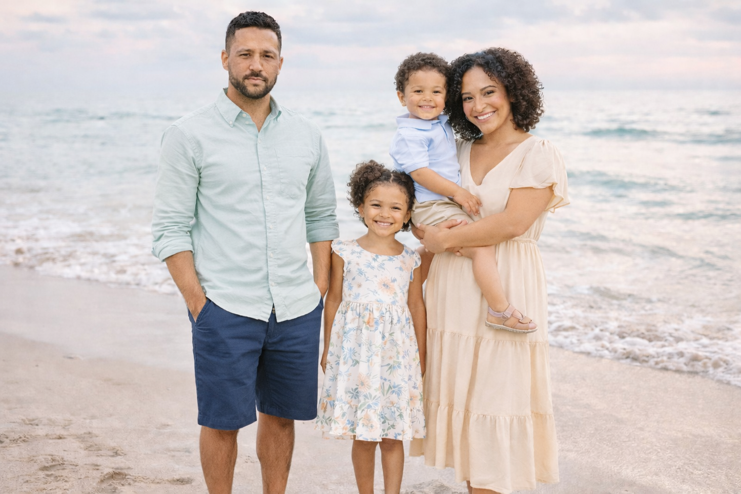 A family wearing coordinated pastel outfits poses together on a sunny beach at sunset.