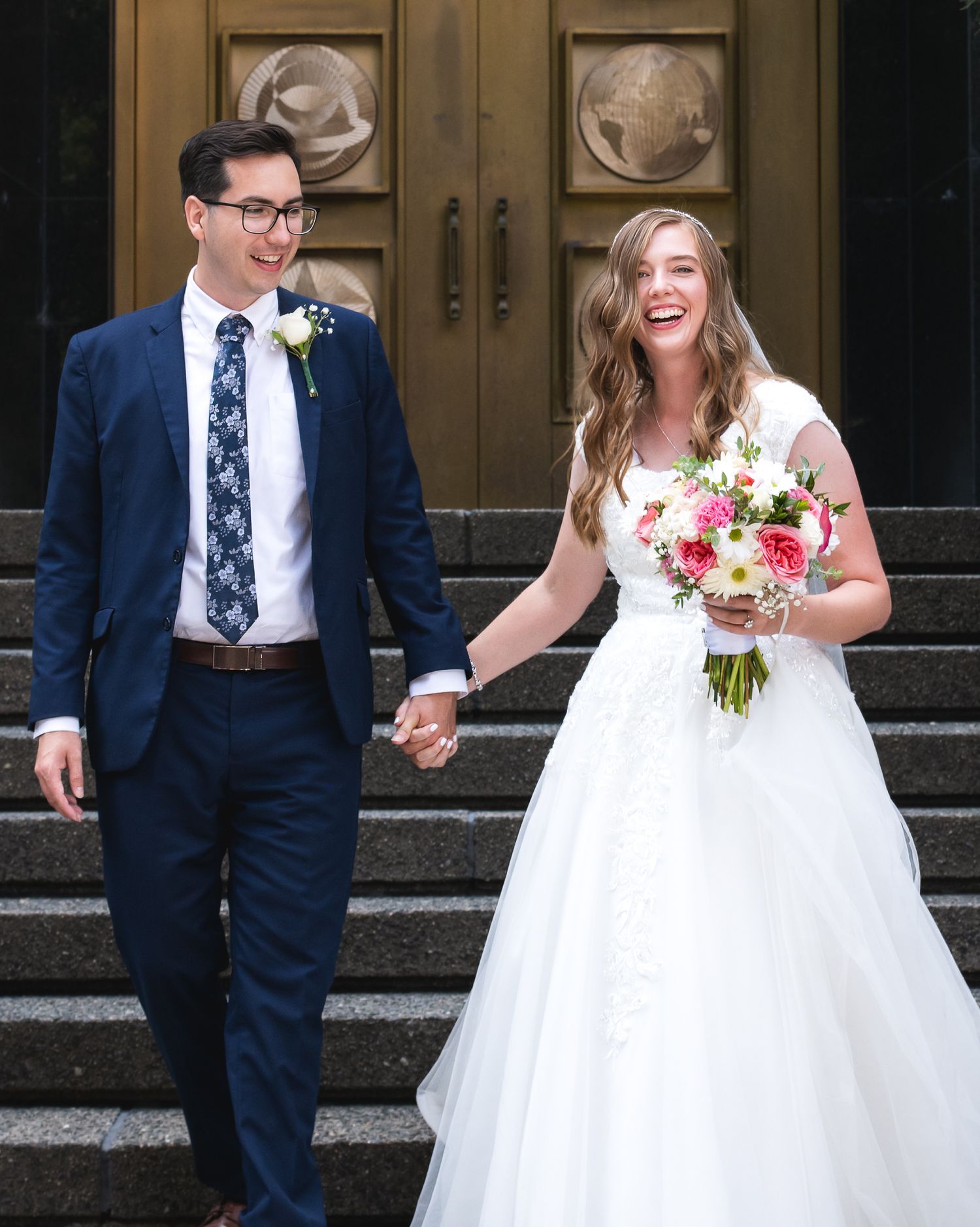 Happy newlyweds walk down steps, bride in white gown holding pink bouquet, groom in navy suit with floral tie.
