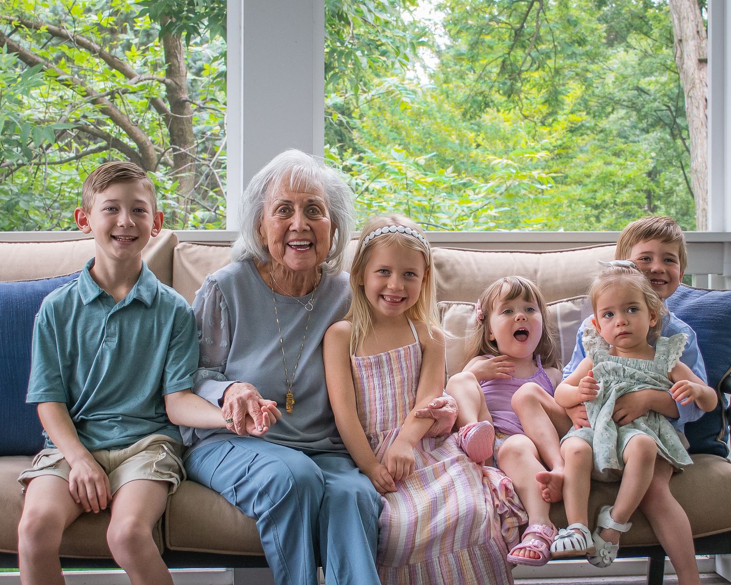 Elderly grandmother smiling on a couch surrounded by five happy grandchildren in a bright sunlit living room.