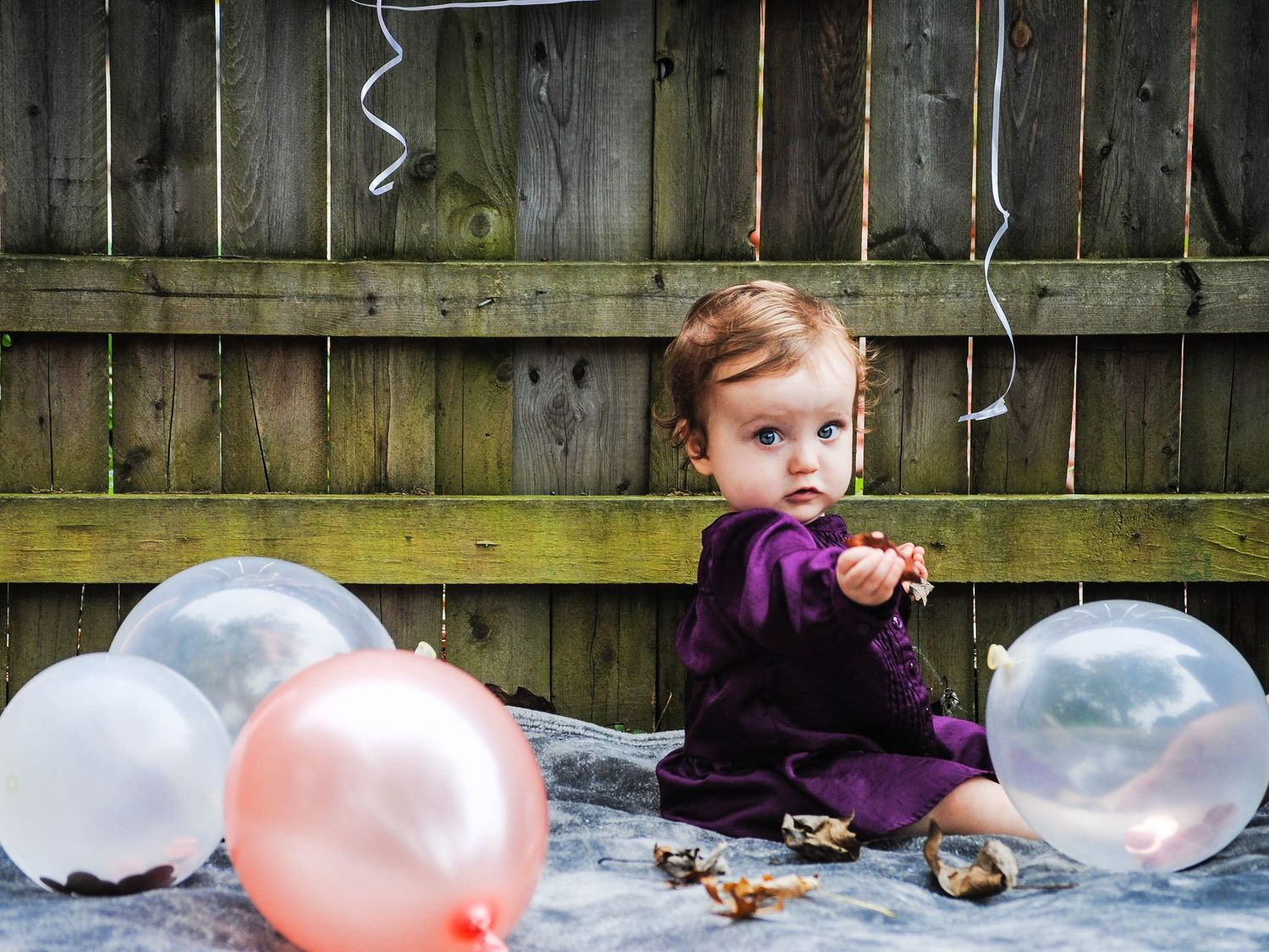 A baby in a purple sweater sits with pink and clear balloons against a wooden fence backdrop.