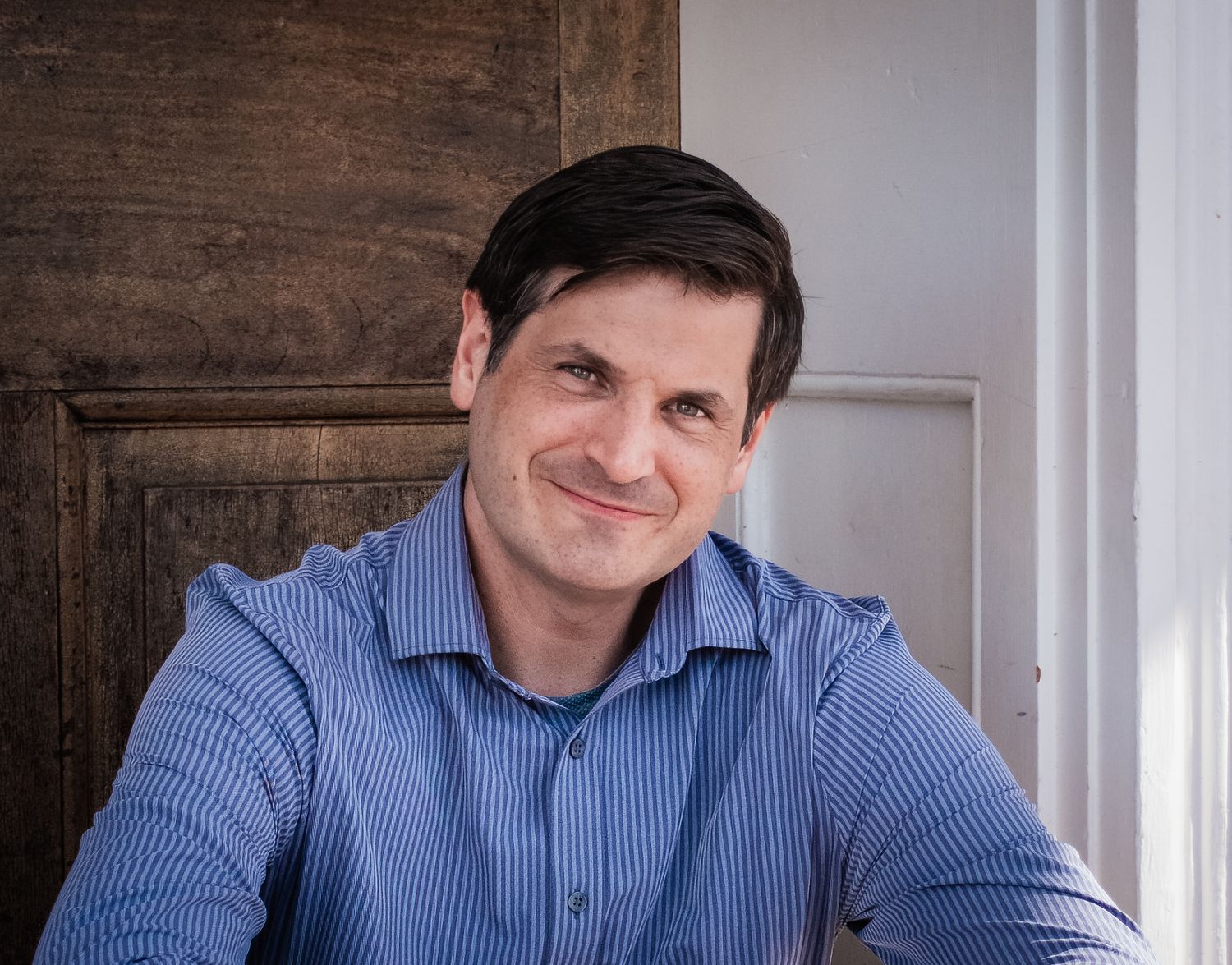 Professional headshot of someone in a blue striped dress shirt smiling warmly against a white and wood paneled wall.