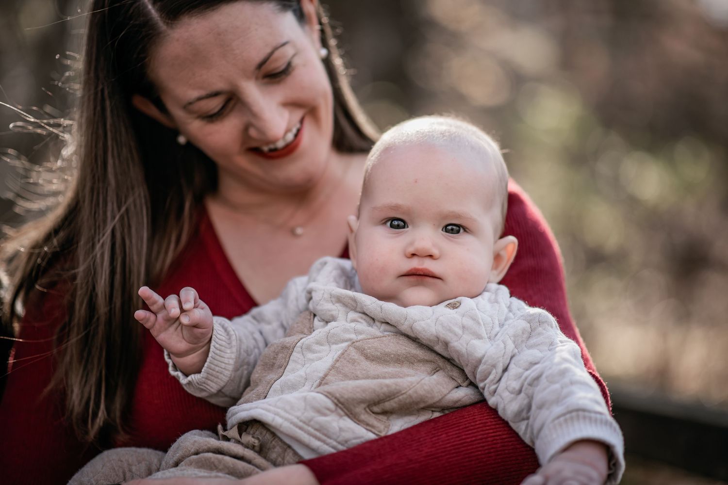 A smiling mother in a red sweater holds her wide-eyed baby dressed in a cozy knit outfit outdoors.