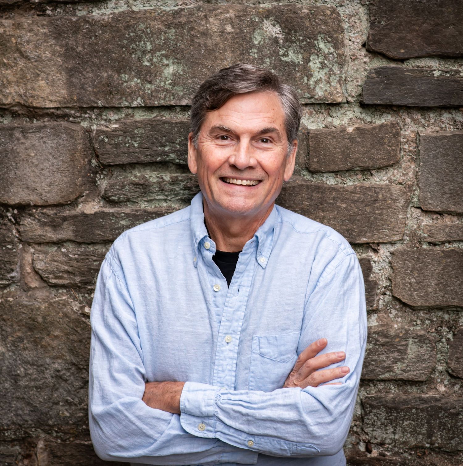 Professional headshot of someone in a light blue button-down shirt posing against a rustic stone wall background.