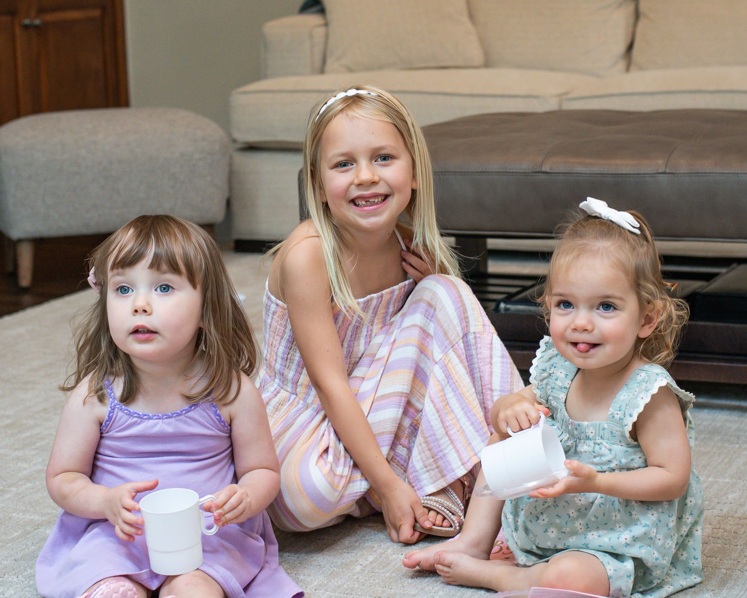 Three young girls sitting on a carpet, smiling and holding white cups in a cozy living room.