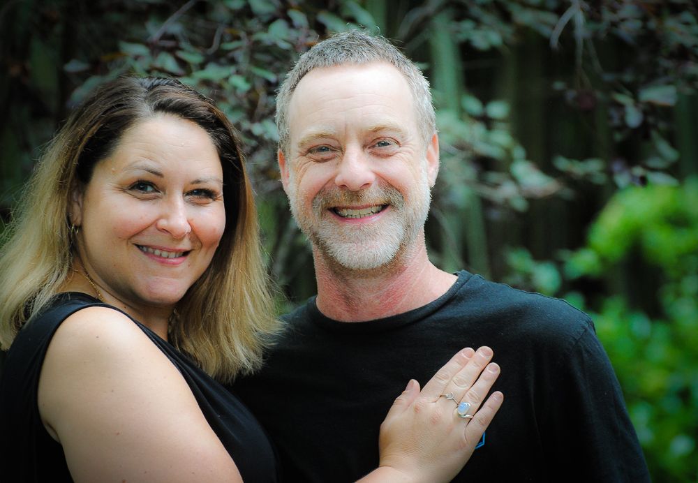 A smiling couple poses outdoors in black tops, with lush green foliage in the background.