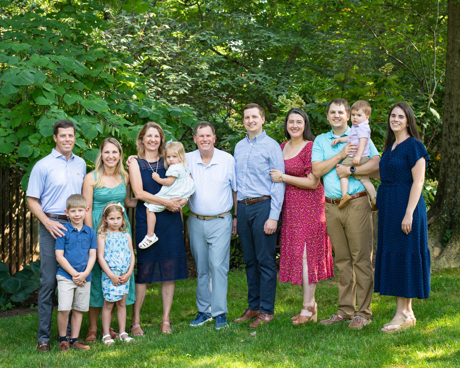 Large extended family group portrait smiling outdoors on green lawn with lush trees in background on sunny day.