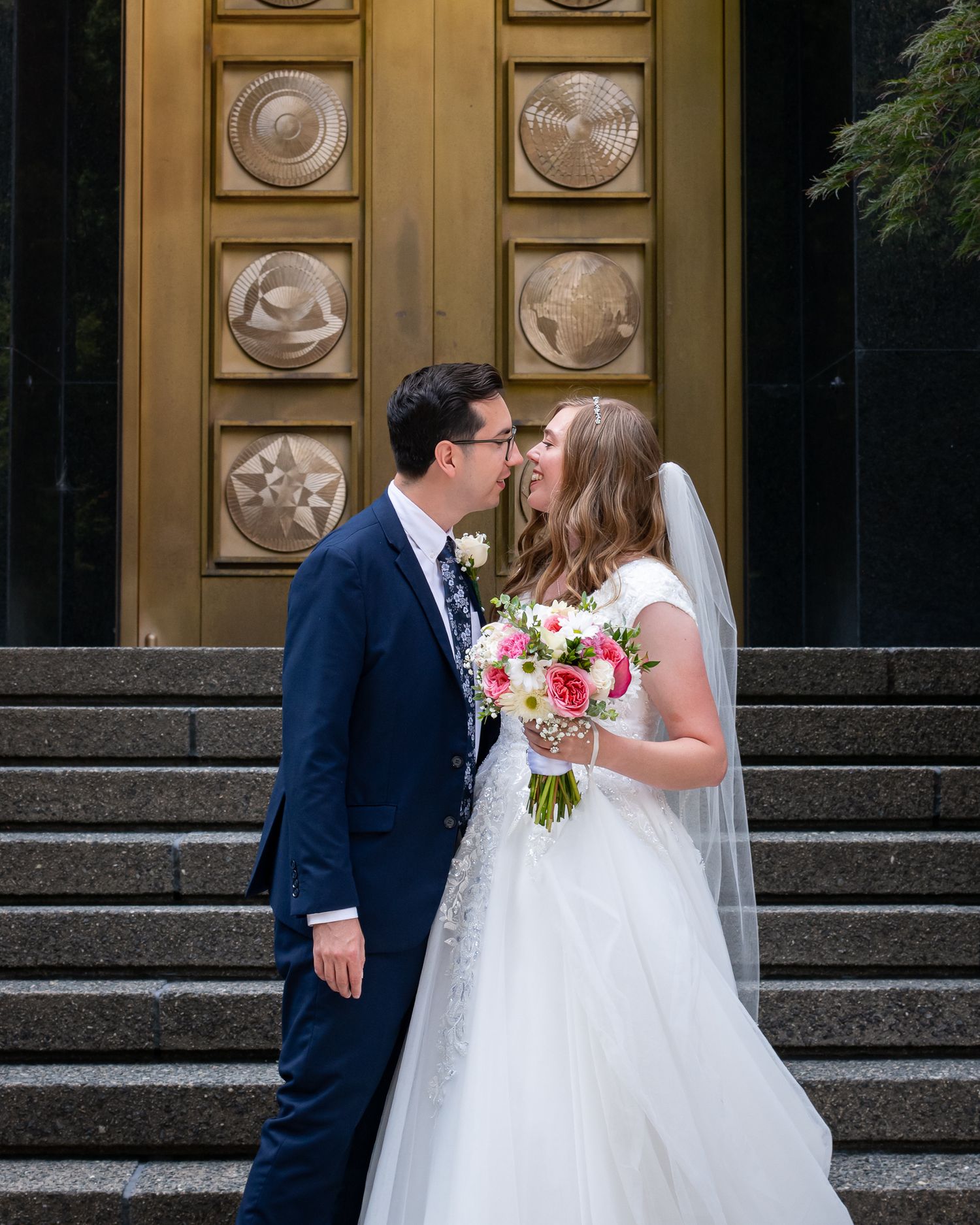 Bride and groom share a kiss on steps before ornate golden doors, bride holding colorful bouquet in white gown.