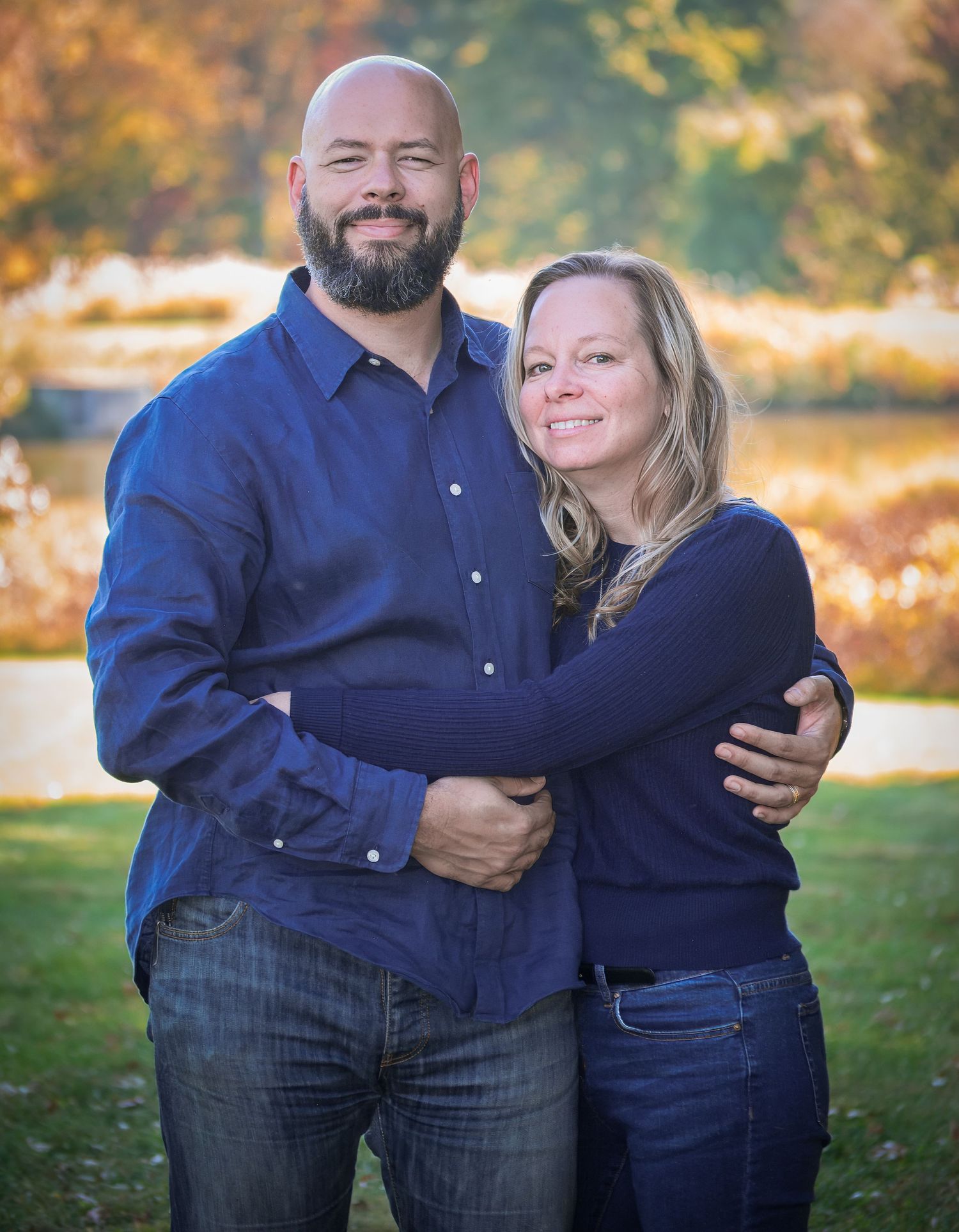 A couple in matching navy blue outfits embrace while standing in a sunlit outdoor setting during autumn.