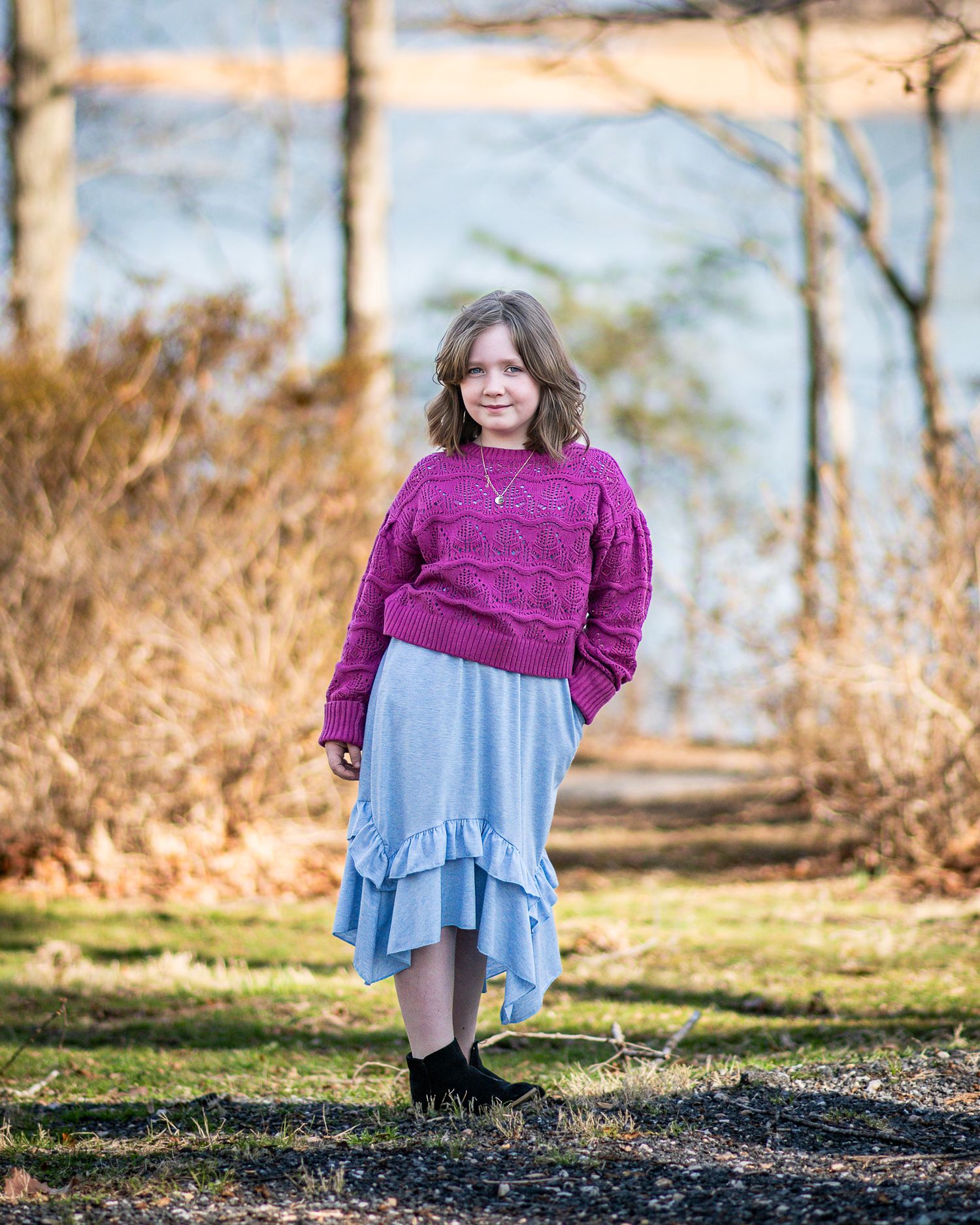 A person wearing a purple sweater and light blue ruffled skirt stands outdoors in a natural wooded setting.