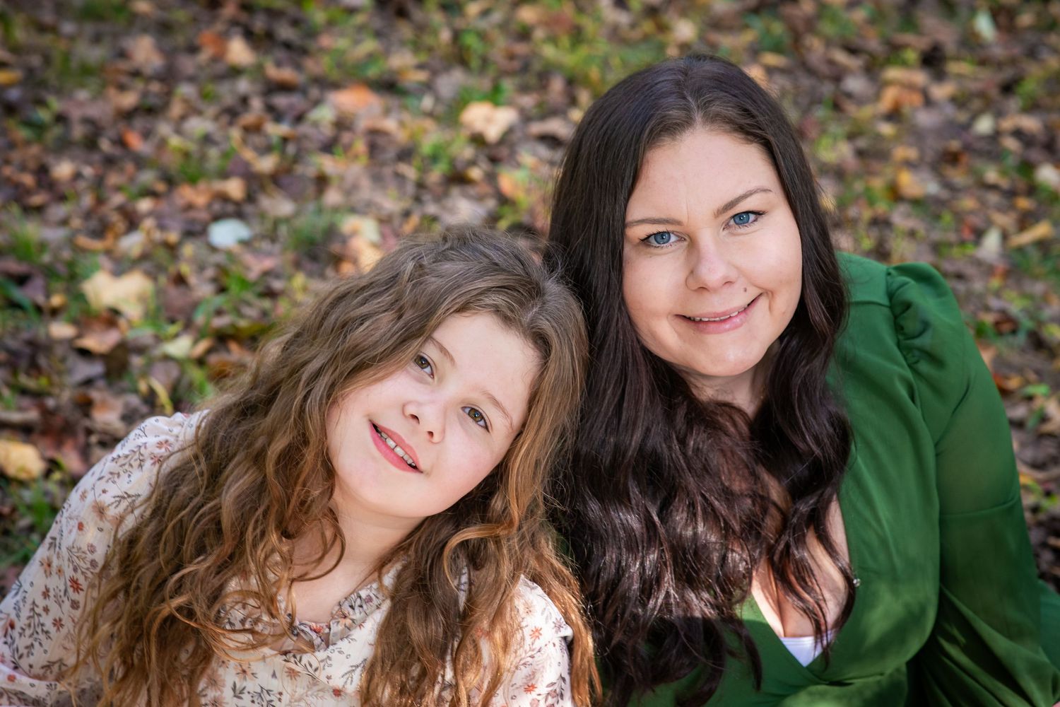 A woman and young girl with curly hair smile together outdoors among autumn leaves.
